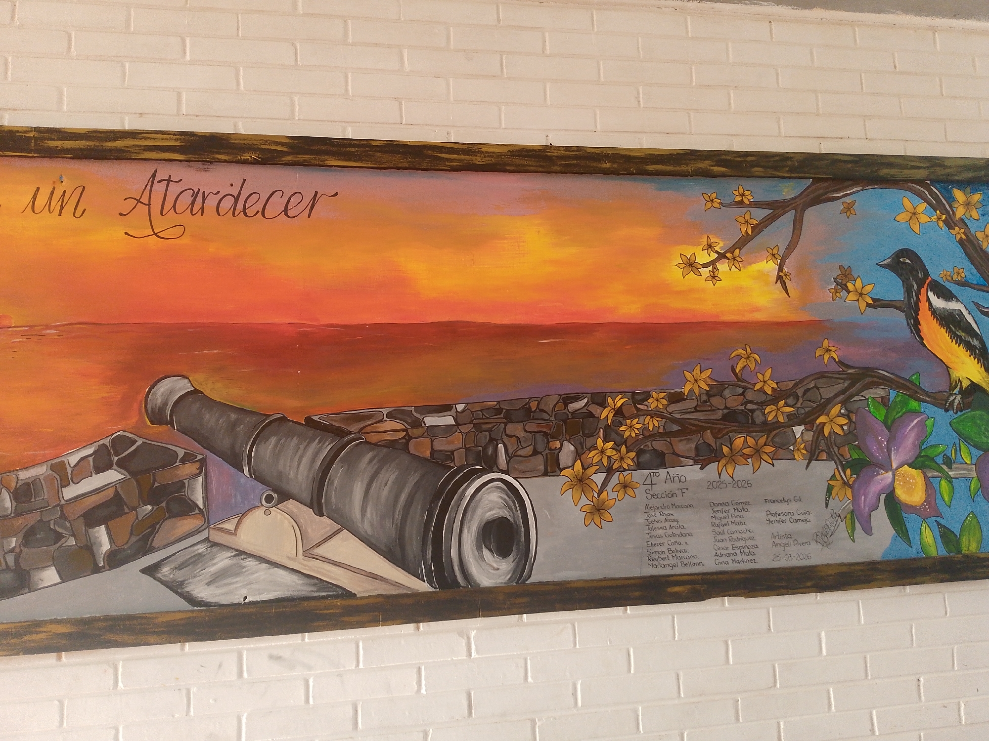The image shows a mural painted on a white brick wall, titled "Un Atardecer" (A Sunset). The painting depicts a sunset over the ocean, with a cannon and stone fortification in the foreground. A tree branch with yellow flowers frames the right side of the mural, with a black and yellow bird perched on it. Purple orchids are also featured on the right side. In the lower right corner, there is text listing names of the students and teachers involved with the mural from 4th year section F during 2025-2026, the artist, and the date 25-03-2026. Juan Griego, Venezuela is not mentioned or inferred from the image.