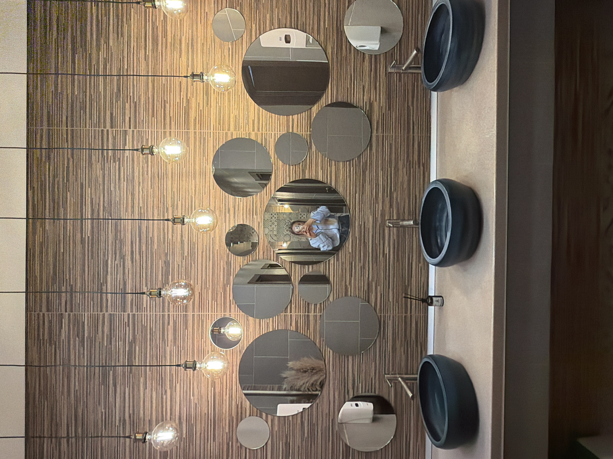 The image depicts the interior of a contemporary public restroom. The precise location within Izhevsk, Russia, cannot be determined from the visual information provided.
The background wall is covered with horizontal rectangular tiles featuring varying shades of brown, creating a textured or wood-like appearance. Above these tiles, a lighter, plain section is visible. Eleven exposed filament light bulbs, each suspended by a black cord, hang from the ceiling at irregular heights across the scene.
Approximately fifteen circular mirrors of diverse diameters are affixed to the tiled wall in an asymmetrical arrangement. These mirrors reflect internal elements of the restroom, including sections of grey tiled walls, a dark brown door, and two white wall-mounted dispensers. One mirror on the left reflects a decorative object resembling dried pampas grass.
A light beige or light brown countertop spans horizontally in the foreground, supporting three dark, oval-shaped vessel sinks. Each sink is equipped with a chrome-finished, single-lever faucet. A small dark bottle containing reed diffusers is positioned on the countertop between the middle and right sinks.
A female individual is visible in the reflection of one of the central mirrors. She is depicted from the upper torso, wearing a light blue button-up shirt over a darker garment, with light-colored hair. She holds a dark smartphone with both hands, positioned to capture a self-portrait (selfie). No other persons or interactions are observed within the frame.