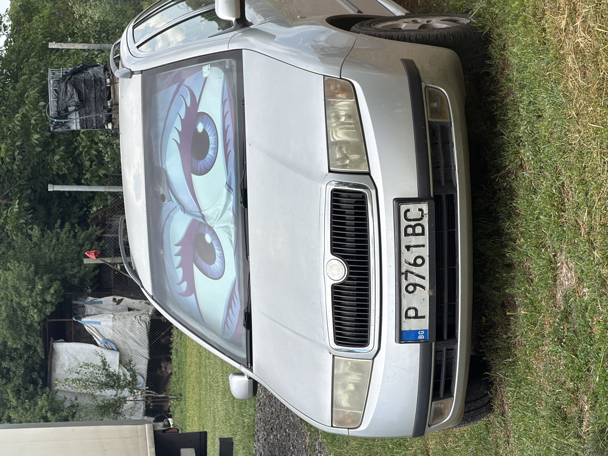 A silver Skoda Fabia station wagon is parked on a grassy area. The car's windshield is covered with a sunshade depicting cartoon eyes. The license plate reads "P 9761 BC" with "BG" in the corner, indicating it is registered in Bulgaria. In the background, there is a chain-link fence, some trees, and what appears to be a shed covered with a white tarp. The setting suggests a rural or semi-rural environment. The image was captured in Chervena Voda, Bulgaria.