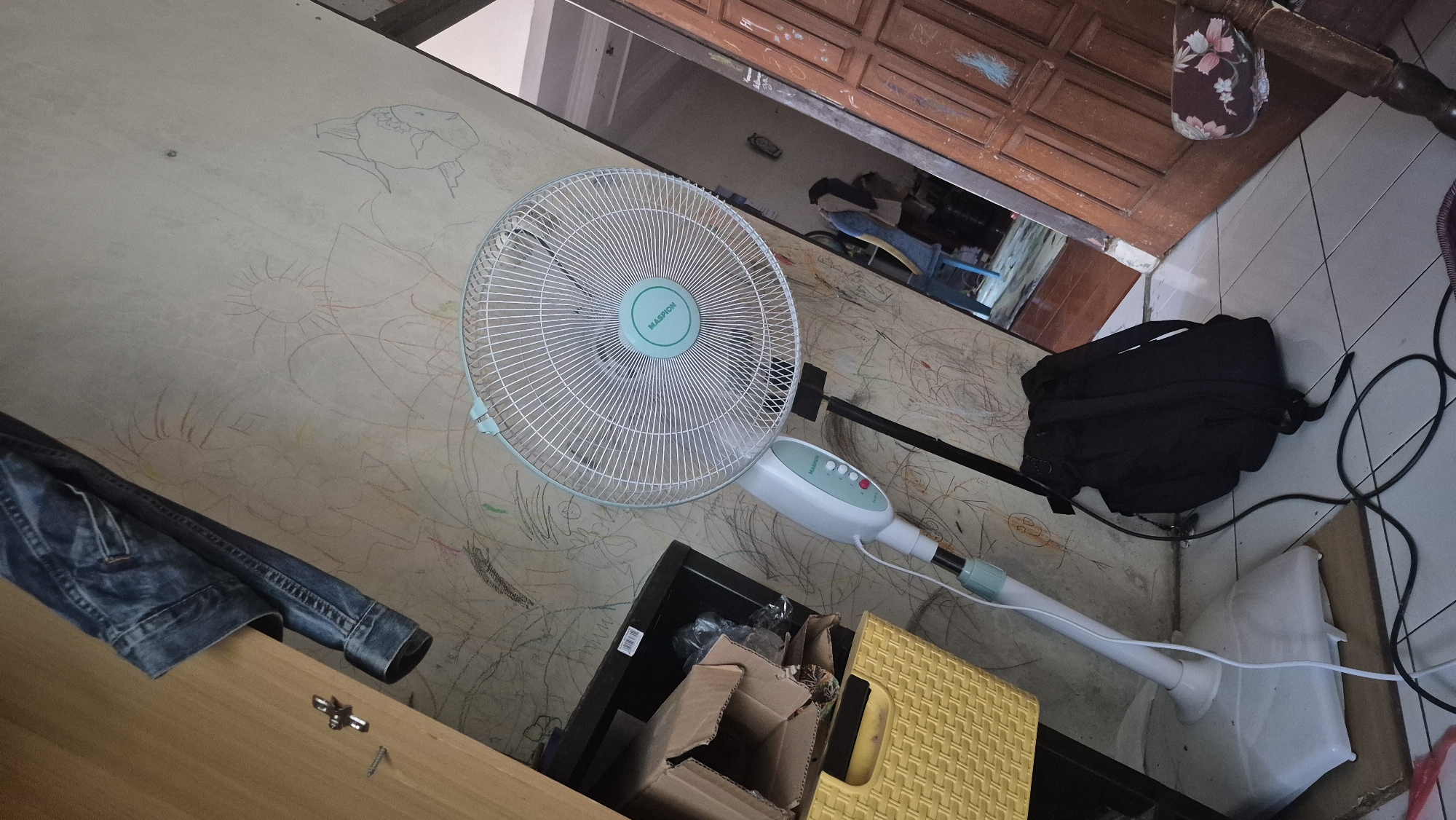 The image depicts a domestic indoor scene, likely a living room or bedroom, with elements suggesting it is located in Limbangan, Indonesia. In the foreground, a white oscillating fan with a teal grill and control panel stands on a tiled floor. A black backpack lies near the fan's base, its power cord trailing. To the left, a yellow woven basket sits next to a cardboard box and a black shelf unit. Partially visible to the far left is a wooden cabinet. The wall behind the fan is covered in what appears to be children's drawings, rendered in various colors. A pair of denim jeans is draped over the cabinet. In the background, a wooden door and a section of a wooden chair are visible, suggesting an adjacent room. The lighting is ambient, and the overall impression is one of a lived-in space.