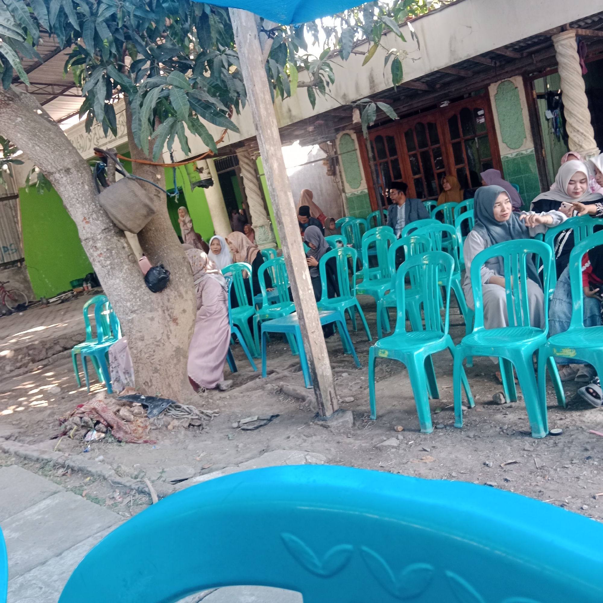 The image depicts an outdoor gathering in Tegowanu Kulon, Indonesia, likely a social event or religious ceremony, indicated by the presence of several individuals dressed in modest attire, many wearing hijabs. The scene is set under a blue tarp, suggesting a temporary shelter, and is partially shaded by the leaves of a large tree with a thick trunk. In the foreground, bright blue plastic chairs are arranged, some occupied by people and others empty.
Several individuals are visible, mostly women, some seated and others standing. One woman in a pink hijab and long dress stands near the tree, while others are seated in the blue chairs. A man in a suit and peci (a type of cap) is seated in the background. The ground appears to be dirt and gravel, with some scattered debris. Behind the gathering, a building with wooden doors and windows, painted green and white, is visible. To the left, a green wall and a bicycle are present, adding to the everyday environment of the location. The overall impression is of a community event taking place in a residential or public outdoor space.