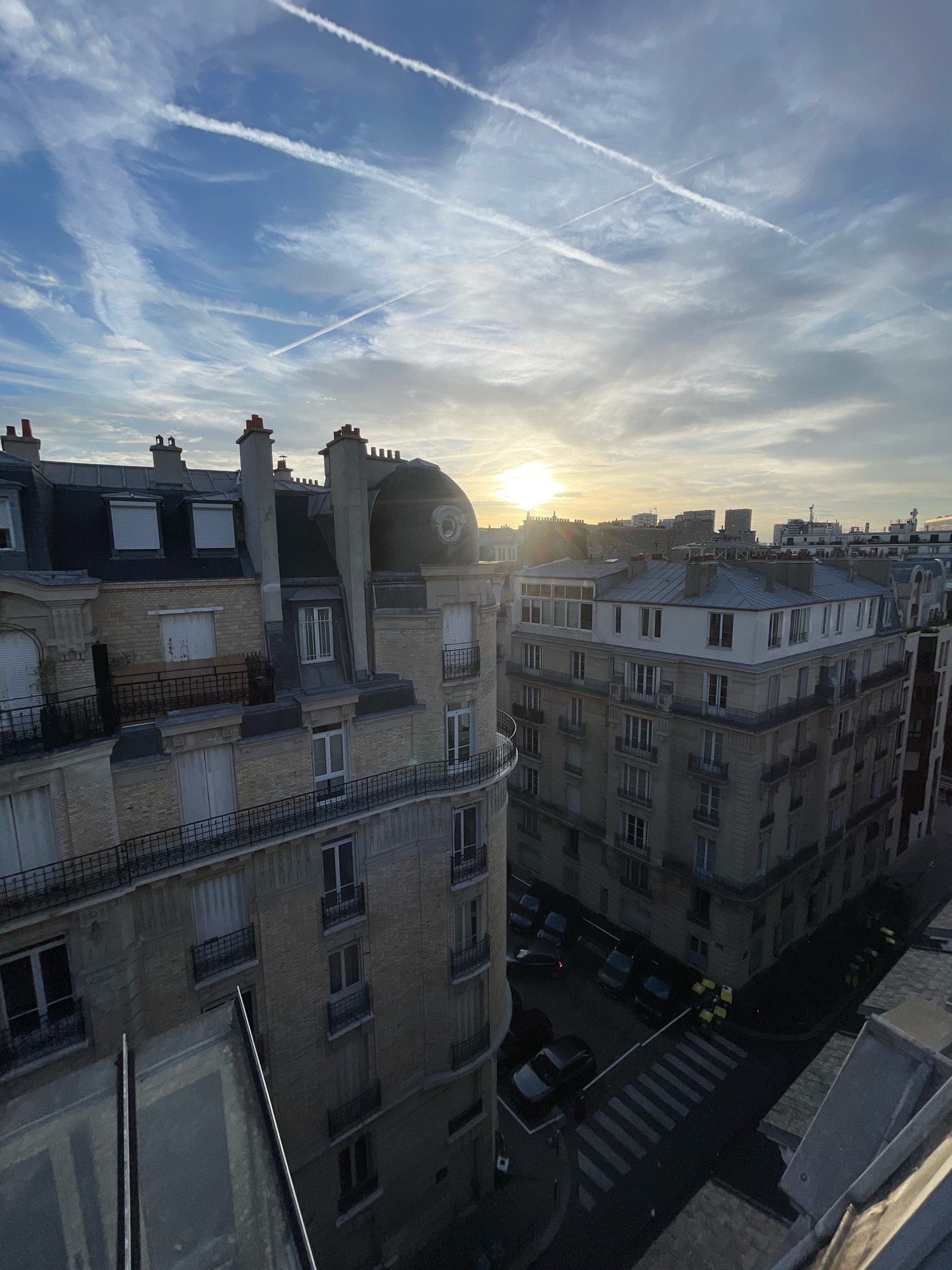 This is a high-angle, full-shot photograph of an urban street scene in Paris, France, during daytime. The scene is dominated by buildings and the sky. The sky is blue with scattered white clouds and contrails from aircraft, suggesting a clear but breezy day. The sun is visible low in the sky, partially obscured by clouds, indicating it is either sunrise or sunset.
The buildings are characteristic of Parisian architecture, featuring Haussmannian style. They are multi-story residential buildings with light-colored stone facades, numerous windows with shutters, and balconies with ornate railings. The rooftops are made of dark gray material, with prominent chimneys and dormer windows. One building on the left has a rounded dome-like structure on its roof.
In the street below, several dark-colored cars are parked or driving. A crosswalk is visible, and several yellow garbage bins are lined up along the sidewalk. The street is paved and appears to be a typical city street.
The foreground includes a portion of a rooftop or balcony railing, made of glass and dark metal, which frames the lower part of the image. There are no people visible in the scene. The overall impression is a quiet moment in a bustling city, captured from a bird's-eye perspective.