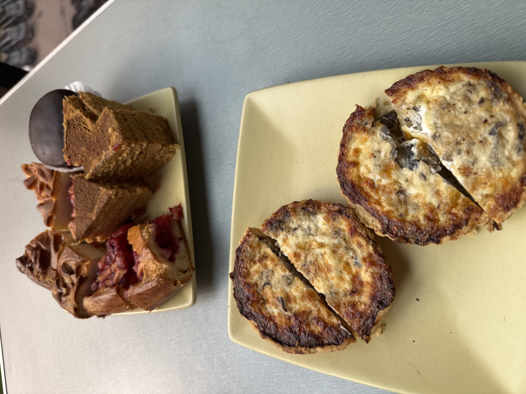 The image presents a top-down view of two plates of food. The plates are pale yellow and square. On the left plate is an assortment of pastries, including pieces of cake, one dark chocolate-covered pastry, and pastries with a red filling, possibly fruit. The pastries are arranged haphazardly. On the right plate, there are two small quiches, each sliced into two halves. The quiches have a browned crust and a filling with dark spots, possibly mushrooms or olives. The background surface is a light gray color. The location of Murino, Russia is extraneous to the visual content of the image.