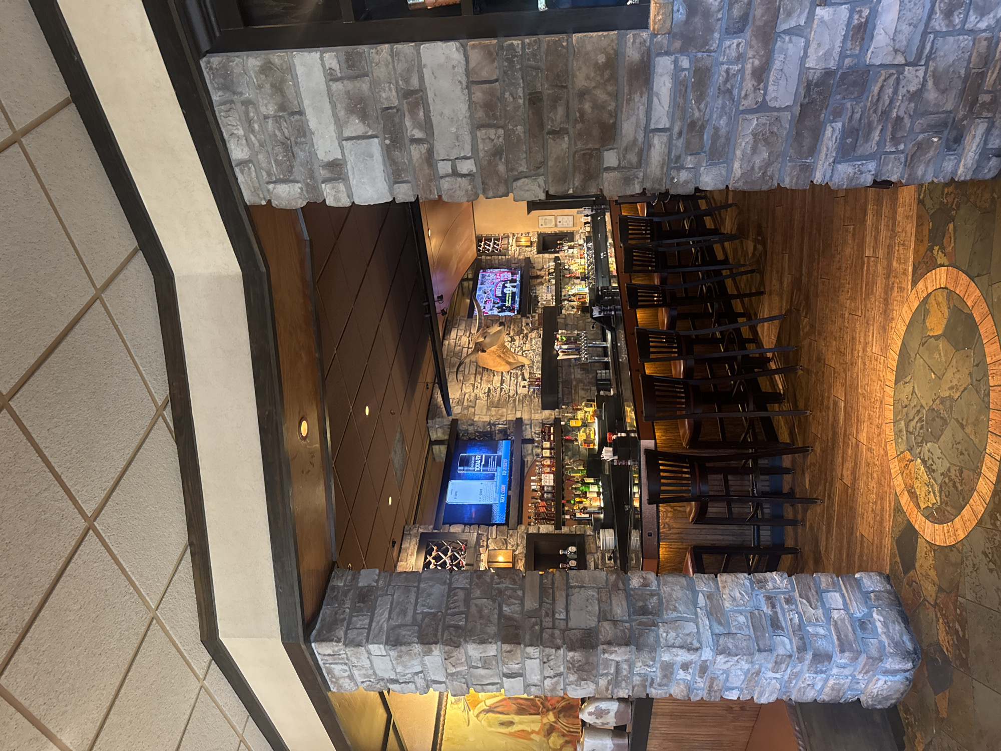 This image captures the interior of a restaurant or bar, taken from an elevated perspective looking down into the main bar area. The scene is framed by heavy stone pillars or arches, made of various shades of gray and brown rough-hewn stones, suggesting a rustic yet upscale ambiance.
The bar itself is well-lit and fully stocked with numerous bottles of liquor and spirits, arranged on shelves behind a dark counter. A series of dark wooden bar stools with slatted backs are neatly lined up along the bar. Above the bar, several flat-screen televisions are mounted. One screen prominently displays a mobile phone interface with text, showing "TEXT CODE TO: 213215" on the right side. Another screen shows what appears to be a sports event, with a figure in a uniform visible. On a wall near the bar, a mounted animal head, possibly an antelope or deer, contributes to the lodge-like decor.
The flooring in the bar area consists of dark wood planks, contrasting with a large circular decorative inlay made of different colored stone or tile, featuring a prominent brick-like ring. To the left, part of the ceiling is visible, made of textured, light-colored square tiles arranged diagonally. The overall setting appears to be a typical American casual dining or bar establishment, likely during off-peak hours as no patrons or staff are present.