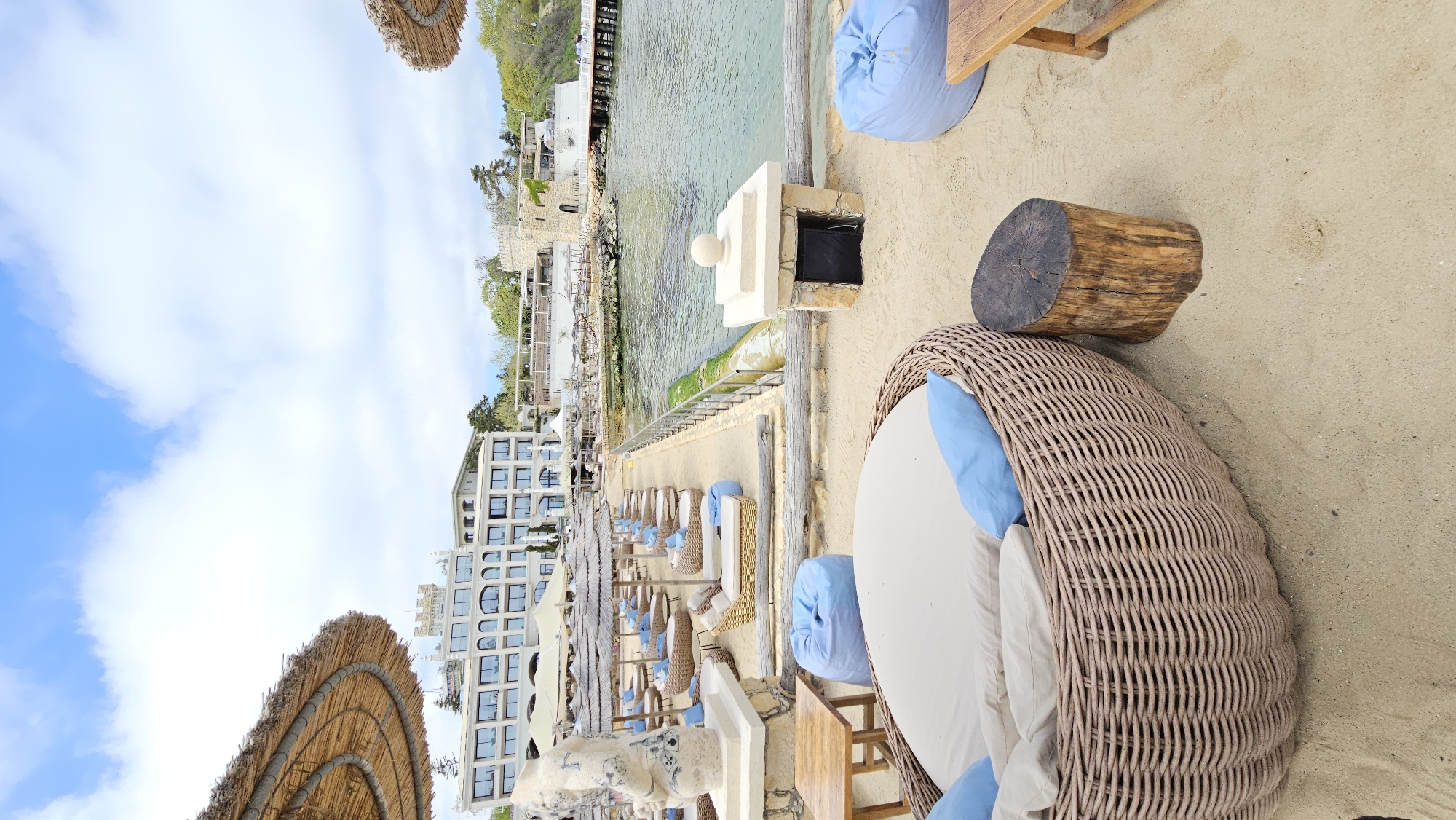 This image depicts a beach resort in Varna, Bulgaria. A wicker chair with blue cushions is situated on the sand. Next to it, a tree stump serves as a makeshift table. A small wooden table with a blue bean bag is nearby. In the background, a coastal hotel and residential buildings are visible alongside a row of beach chairs with thatched umbrellas. The sea is clear and calm. The sky is partly cloudy.