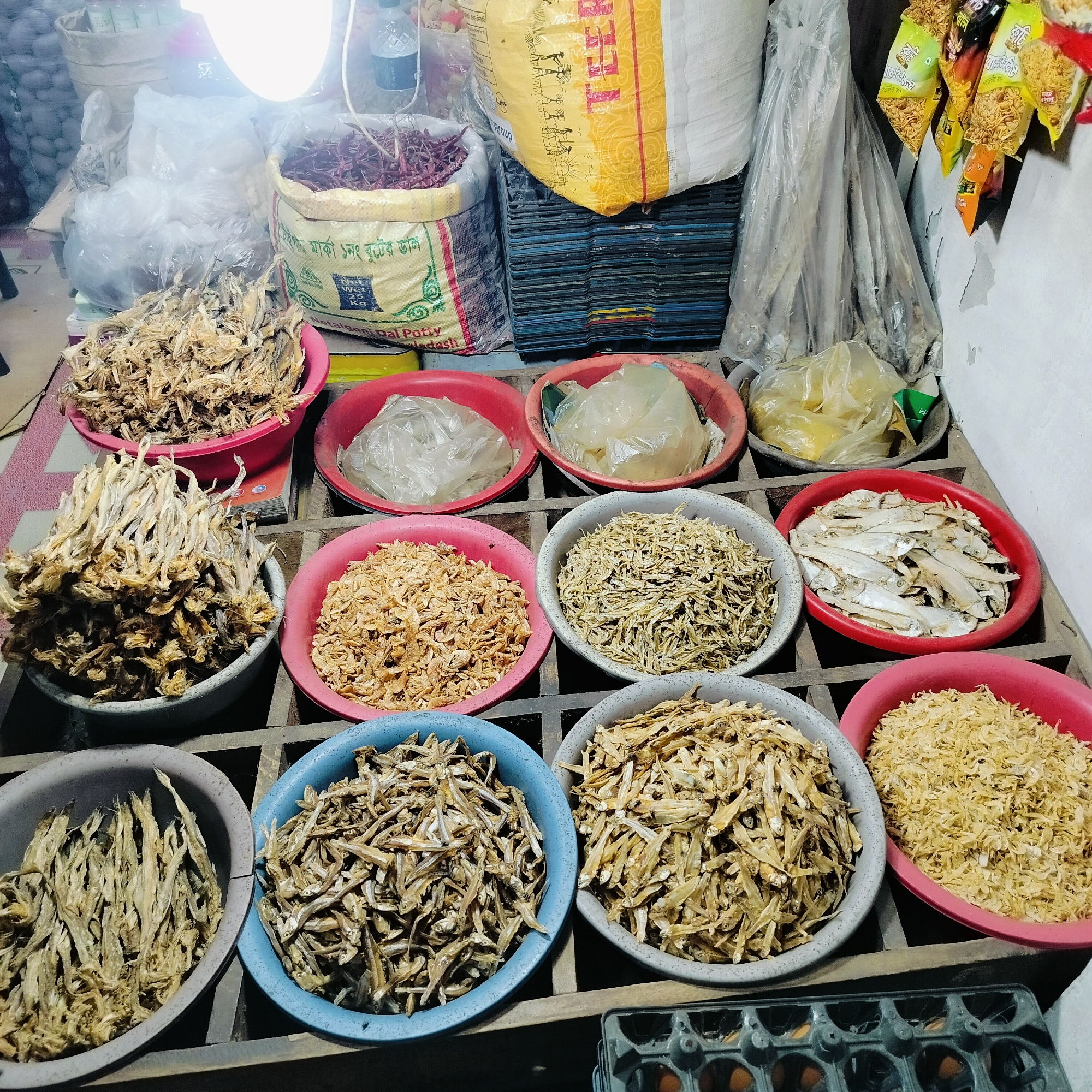 The image depicts a vendor's display of dried fish in a market in Dhaka, Bangladesh. Bowls of various sizes are filled with dried fish of different types, colors, and sizes, arranged on a wooden shelf. Some bowls are made of gray plastic, while others are made of red or blue plastic. The shelf has a grid-like structure and sits atop what appears to be an egg carton and possibly other items. Above the dried fish, bags of dried goods, spices, and other items can be seen. The scene is lit by an overhead light, causing glare on some of the items. The market appears to be busy with activity.