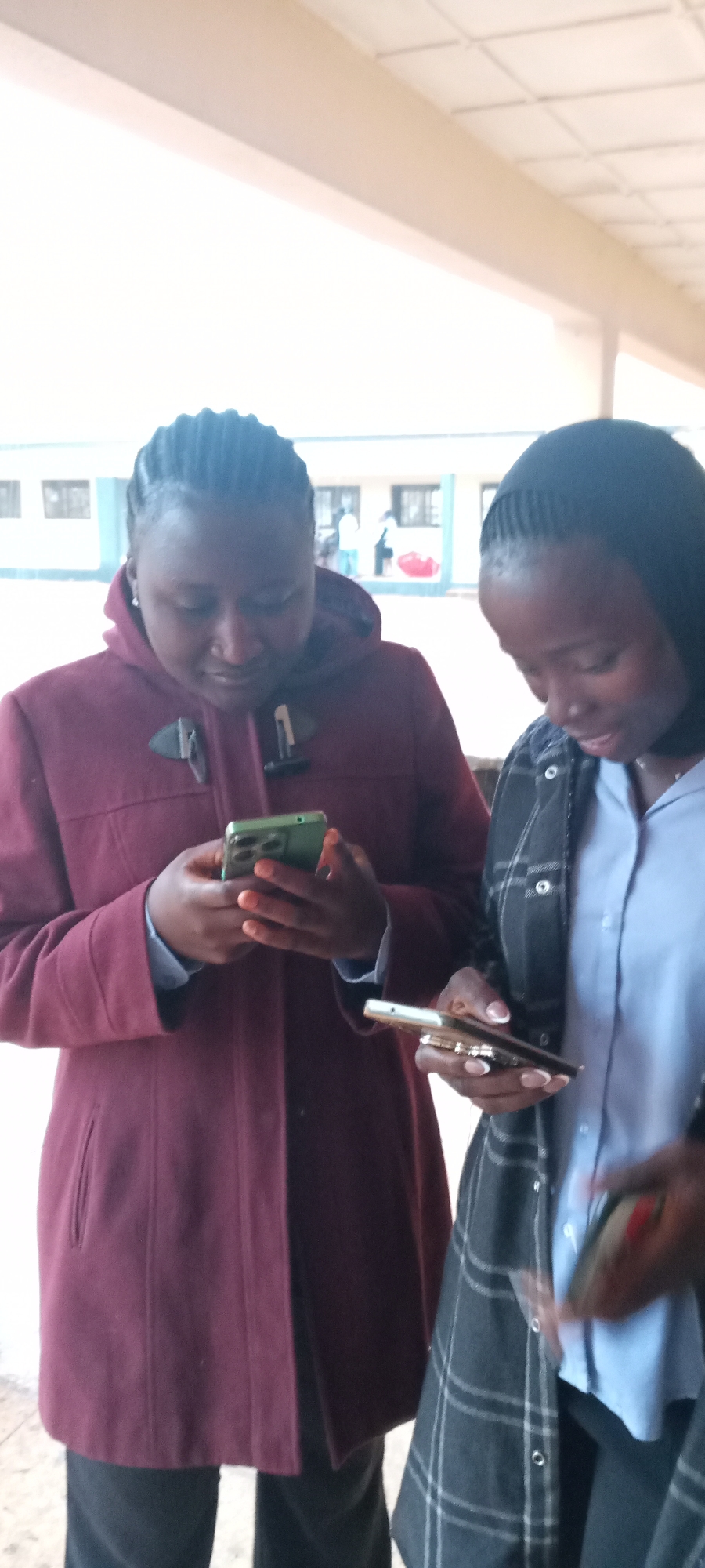 Two young women stand close together, each looking down at a smartphone in their hands. The woman on the left wears a maroon hooded coat and the woman on the right wears a blue button-down shirt with a grey plaid jacket. They appear to be in an outdoor covered area, possibly a schoolyard or similar location, as a building with windows is visible in the background. The city and location are unknown.