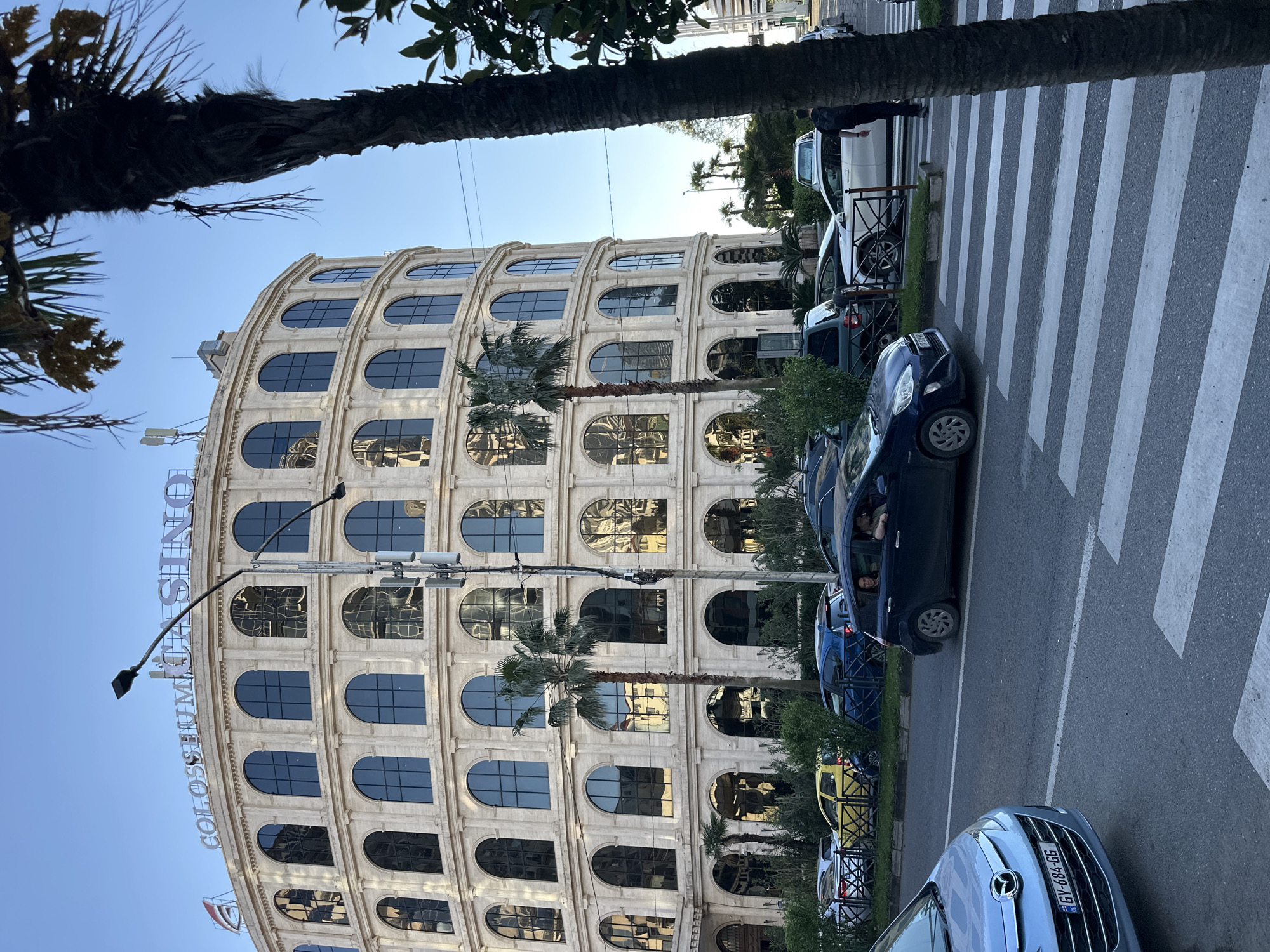 The image captures a street scene in Batumi, Georgia. In the background is a large, curved building with multiple arched windows reminiscent of the Roman Colosseum. The building is labeled "COLOSSEUM CASINO". Several palm trees are planted along the street. A crosswalk is visible in the foreground. Several cars are parked along the street, and one dark-colored car has at least two occupants visible inside. A lamppost is positioned near the building. The sky is clear and blue.