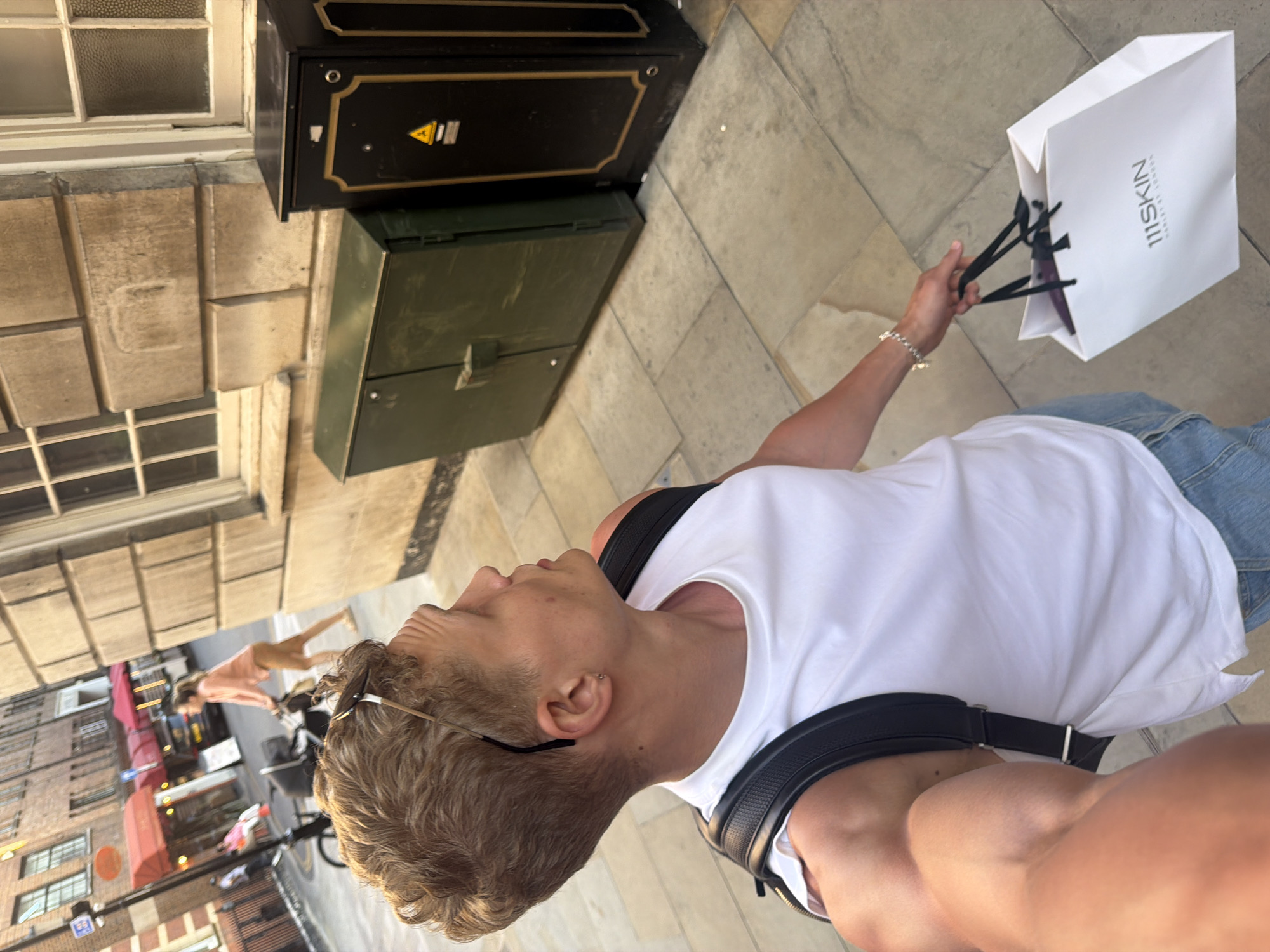 A medium shot captures a person holding a white shopping bag with the logo "111SKIN HARLEY STREET LONDON." The person wears a white t-shirt, blue jeans, and a black backpack, with sunglasses perched on their head. A bracelet adorns their left wrist. To the left, an electrical box sits next to a building with stone facade. In the background, a street with storefronts and a cyclist are visible. The scene is taken on the streets of London, United Kingdom, with apparent sunshine.