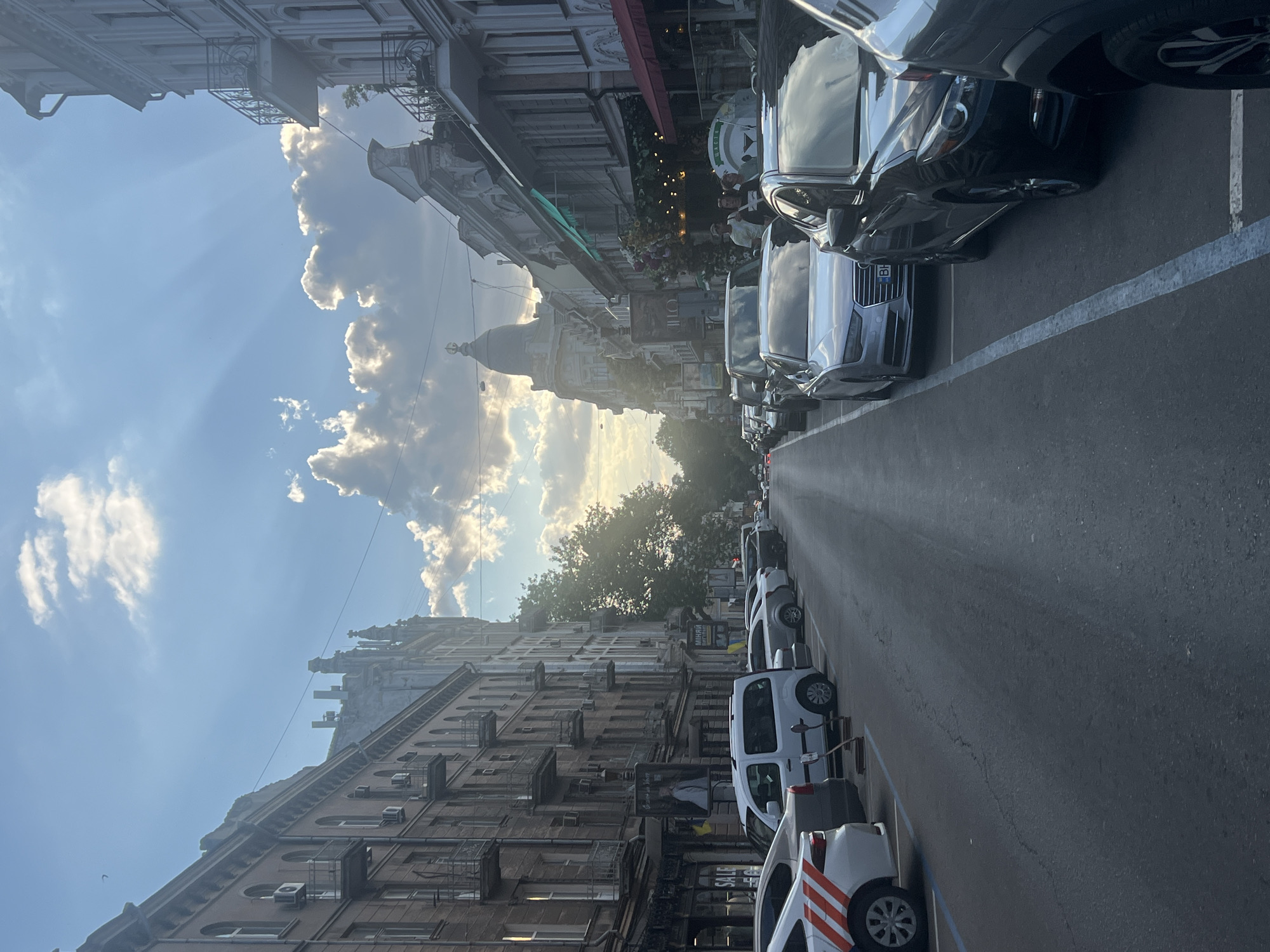In Odesa, Ukraine, a street view captures buildings lining both sides of a road filled with parked cars. Sunlight beams through dramatic clouds in a bright blue sky. The architecture of the buildings displays ornate details and balconies. Several vehicles, including a white van and various sedans, are parked along the street. People can be seen walking near some of the buildings and in the background. The scene suggests a typical urban environment with a notable sky display.