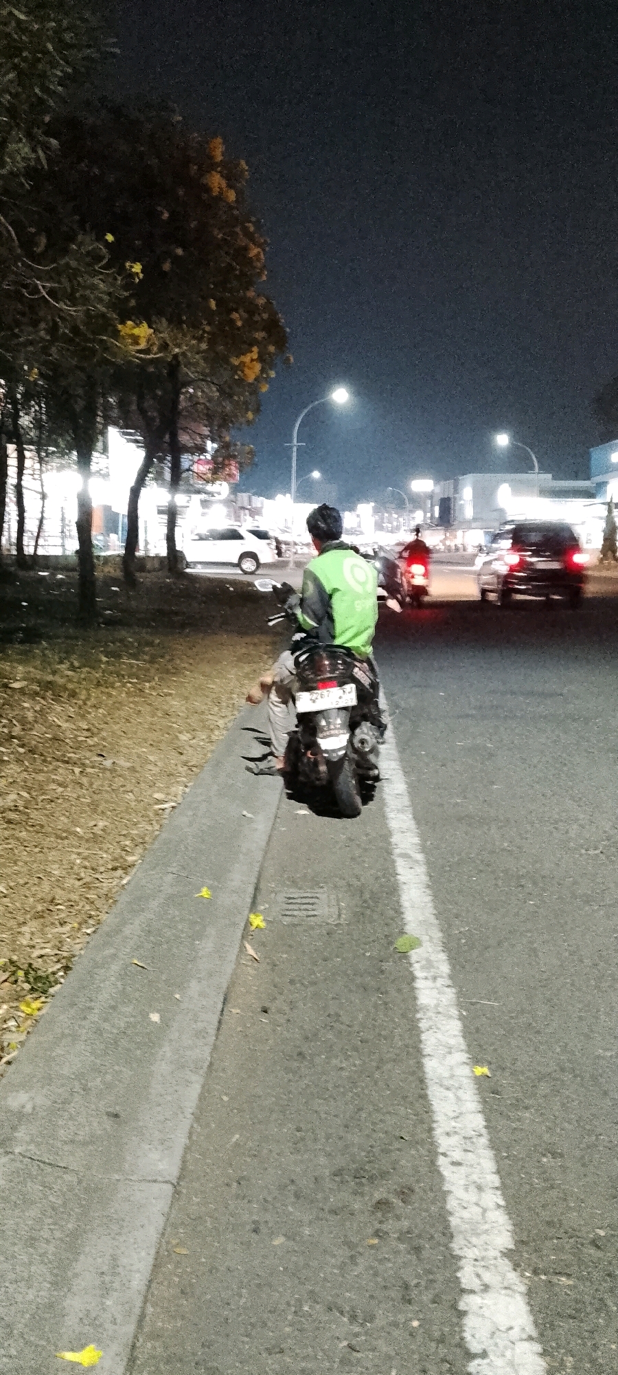 A person wearing a bright green jacket with a circular white logo on the back sits astride a dark-colored motorcycle on a paved road at night. The motorcycle is parked on the right side of the road, partially on the shoulder and partially in the traffic lane, marked by a white line. The rider is facing away from the camera, looking towards the left of the frame. Their legs are visible, wearing light-colored pants and sandals. The motorcycle has a license plate that reads "F 2267 SJ" and has a red taillight illuminated.
The environment appears to be an urban or suburban area in Cileungsi, Indonesia. Trees with sparse foliage and some yellow flowers line the left side of the road, casting shadows. In the background, several buildings with illuminated windows are visible, as well as streetlights and other vehicles on the road. A white car is parked on the left side, and other cars, including a dark-colored SUV with illuminated taillights, are driving on the road. The sky is dark, indicating it is nighttime. The road surface is asphalt, and a concrete curb separates the road from the grassy and leaf-strewn shoulder.
