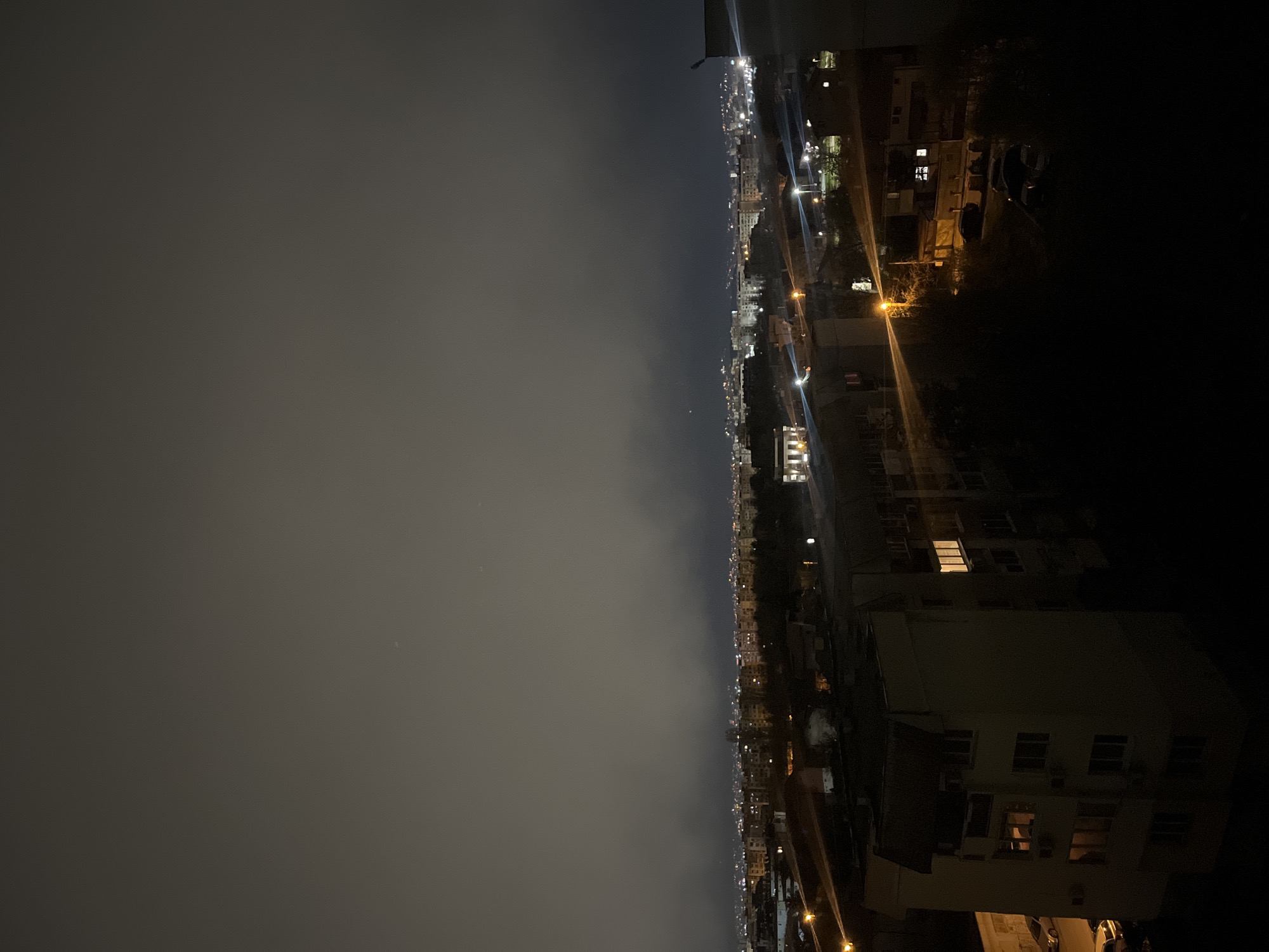 Here's a detailed description of the image:
**Location:** The image appears to be taken from a high vantage point, likely a rooftop or high-rise building in Sofia, Bulgaria, judging by the urban skyline.
**Environment:** The scene depicts a nighttime cityscape. The sky is overcast and shrouded in low clouds or fog.
**Subjects and Objects:**
* **Cityscape:** A long stretch of urban landscape is visible, extending into the distance.
* **Buildings:** A mix of residential and commercial buildings of varying heights.
* **Lights:** Streetlights create lines of illumination along the roads. Apartment windows emit light.
* **Roads:** There are roads that are identifiable due to the car and streetlights.
* **Foreground:** A dark area occupies a substantial portion of the frame. This area includes the rooftops of buildings close to the camera.
**Actions and Interactions:** No active actions or interactions of people can be identified. The scene appears still and quiet.