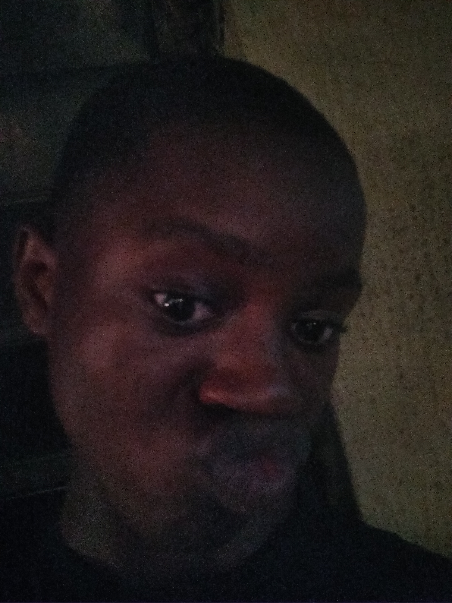 A dimly lit, grainy close-up portrait features an individual, likely male, with dark skin and short hair, looking directly at the camera. The subject's face fills much of the frame, illuminated by a direct, low light source, evident from distinct reflections in both pupils. The expression appears casual, possibly with a slight pout or relaxed demeanor. The background is dark and features a textured surface, resembling a wall, in muted, earthy tones, indicating an indoor setting. The low light conditions contribute to the image's significant grain and reduced clarity. No other objects or visible text are discernible. The image likely depicts a person in Shangisha, Nigeria.