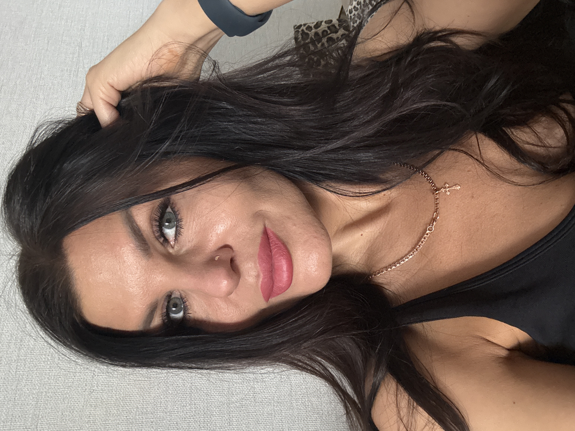 A woman with long, dark hair is lying down, facing the camera. Her eyes are gray. She is wearing a rose gold necklace with a small cross pendant. She has a fitness tracker on her wrist. She is wearing a black tank top. The background is a light gray surface. No information regarding Chaikovsky, Russia, is discernible in the image.
