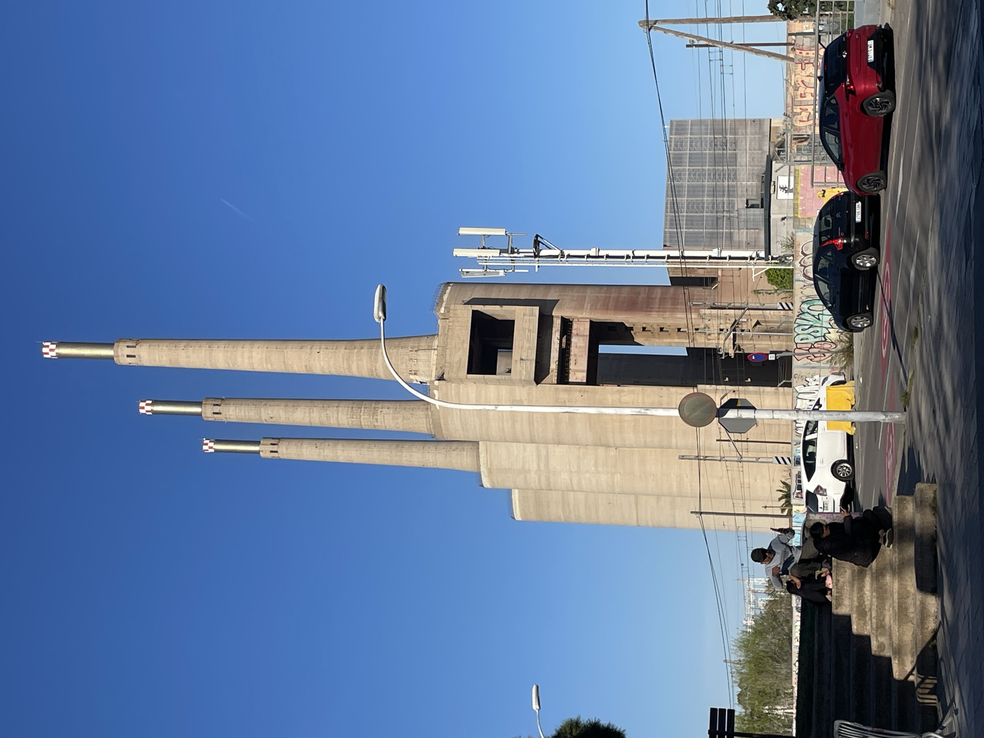 The image depicts the Three Chimneys of Sant Adrià de Besòs, Spain, a prominent industrial landmark. The three tall, concrete chimneys dominate the left side of the frame, reaching towards a clear blue sky. A large, box-shaped structure, also made of concrete, is connected to the base of the chimneys. Overhead power lines and lighting fixtures are visible near the towers. On the right, the image shows a street scene with several parked cars, including a red one and a black one. A small group of people is gathered on some steps, seemingly engaged in a conversation. A white car is parked nearby, partially obscured. In the background, there is a grid-like metal structure. Graffiti is noticeable on some of the structures near the cars. The scene is well-lit, suggesting it is daytime.
