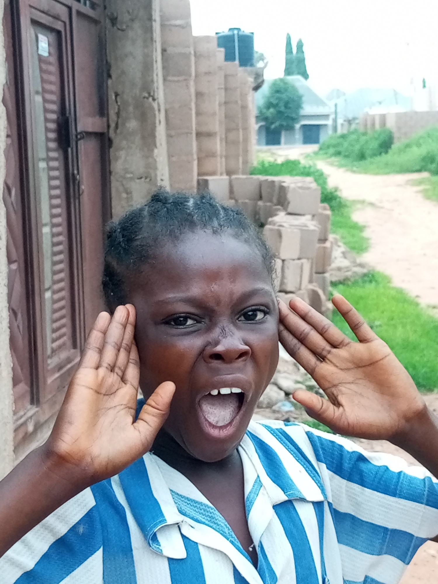 A young girl with dark skin and braided hair is pictured in a close-up shot, looking directly at the camera with her mouth open and hands cupped around her ears as if shouting or listening intently. She is wearing a blue and white striped shirt. The background shows a partially constructed building with bricks stacked nearby, and beyond that, a dirt road flanked by green vegetation and some distant structures. The overall setting suggests an outdoor environment in a developing area, possibly a residential or construction site in Tempure, Nigeria.