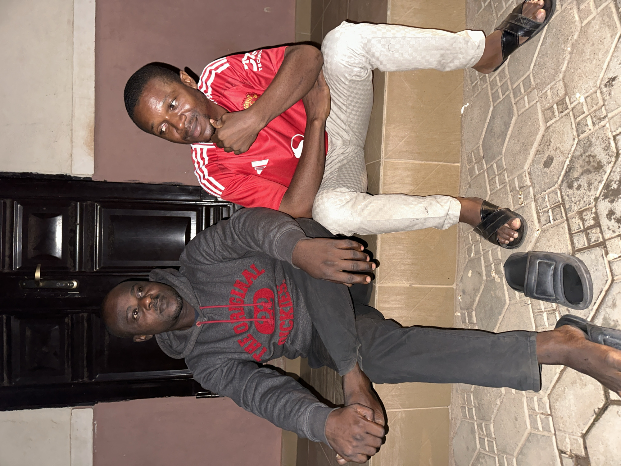 Two men are sitting on a tiled step in front of a dark wooden door. The man on the left is wearing a grey hooded sweatshirt with red lettering and black pants. He is looking off to the side with his legs crossed. The man on the right is wearing a red shirt with white stripes on the sleeves and light-colored pants. He is looking at the man on the left and has his chin resting on his hand. The ground in front of them is also tiled with a geometric pattern. The environment appears to be an outdoor setting, possibly in the city of Narayi, Nigeria, given the architectural style and the appearance of the individuals.