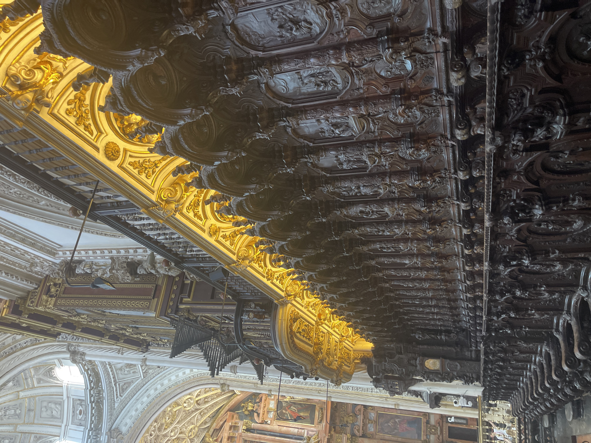 The image captures the ornate wooden choir stalls within the Great Mosque-Cathedral of Córdoba, Spain. The foreground is dominated by the intricately carved dark wood of the choir stalls, featuring detailed relief sculptures depicting biblical scenes and figures. Above the stalls, a richly gilded ceiling with elaborate decorative molding is illuminated by warm light. The background reveals the grandeur of the cathedral's architecture, with high, arched ceilings, whitewashed walls, and a glimpse of a large dome with arched windows. The perspective is angled upwards, emphasizing the scale and artistry of the interior. No people are clearly visible in the main portion of the image, though a distant figure can be seen on the left.