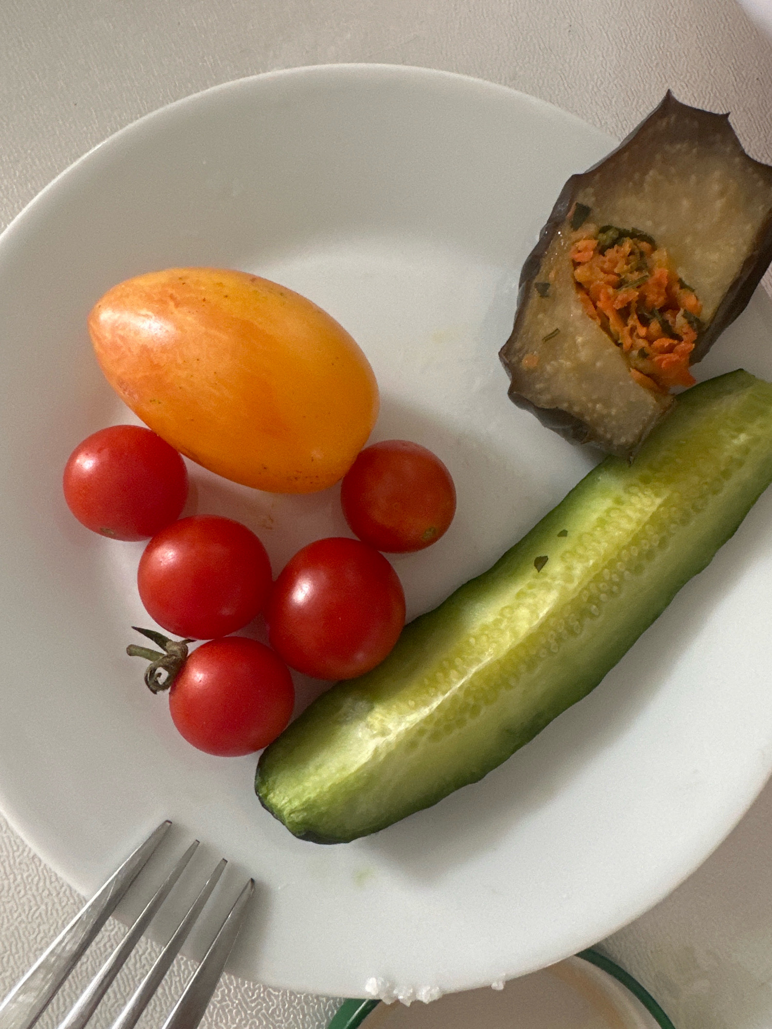 A white plate holds a collection of fresh vegetables and a piece of stuffed eggplant. On the left side of the plate are five cherry tomatoes, two of which are clustered together at the bottom, with one visibly attached to a vine. Above these, an oblong, orange tomato rests. In the center of the plate, a long, halved cucumber lies horizontally, its green skin and pale, seeded interior clearly visible. To the right, a slice of dark purple eggplant is stuffed with a mixture of finely chopped orange vegetables, possibly carrots, and green herbs. A fork with four tines is positioned at the bottom left corner of the plate. The entire arrangement is set against a light-colored, textured surface, possibly a tablecloth or placemat. There is no indication of a specific location in Nevinnomyssk, Russia.