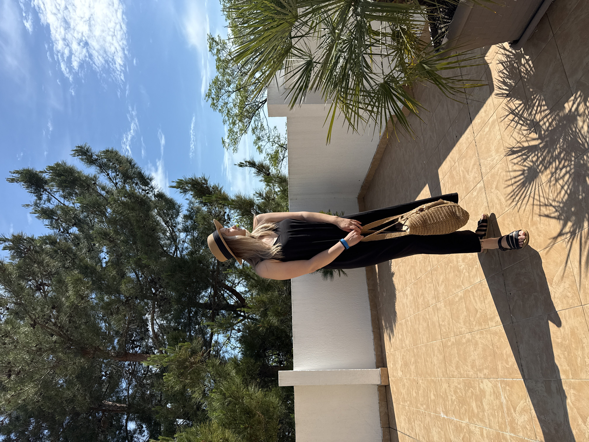A woman stands on a tiled balcony, looking to her right, with a straw hat on her head and a black dress. She holds a woven bag in her right hand and wears black sandals. A blue bracelet is visible on her left wrist. Behind her, a white wall and railing extend across the balcony, with a potted palm tree on the right side. Pine trees and a bright blue sky with scattered clouds form the background. The scene is illuminated by sunlight, casting shadows on the balcony floor. The location is the city of Nebug, Russia.
