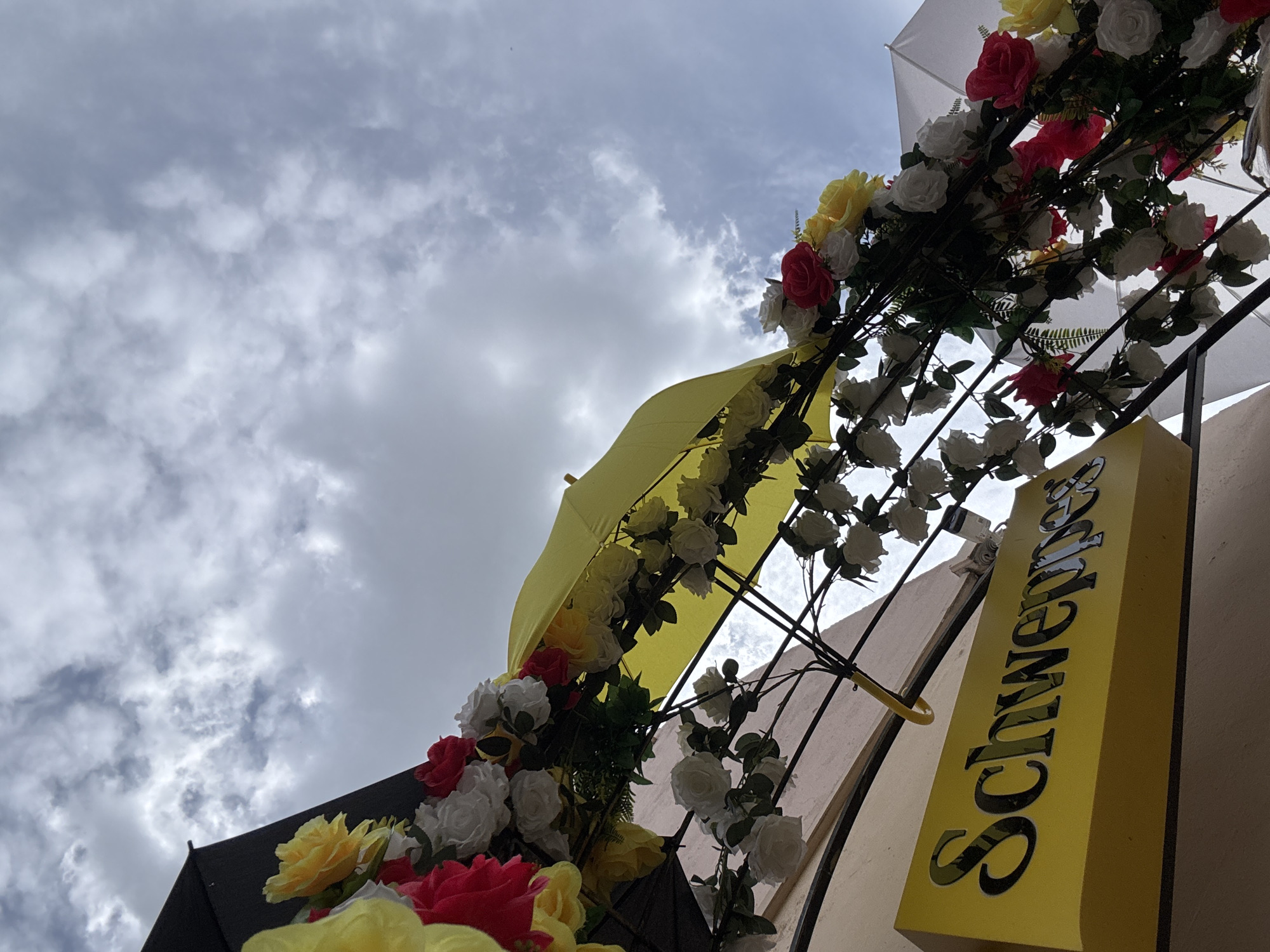 The image shows an outdoor scene with a cloudy sky. In the foreground, a yellow sign with the text "Schweppes" is visible. Above the sign, an archway decorated with artificial flowers and umbrellas is erected. The flowers are in red, white, and yellow colors. Two open umbrellas, one yellow and one black, are part of the archway. There are also other white umbrellas partially visible in the background. The overall scene suggests a festive or decorative setup, possibly for an event or as part of street decoration in an urban environment.