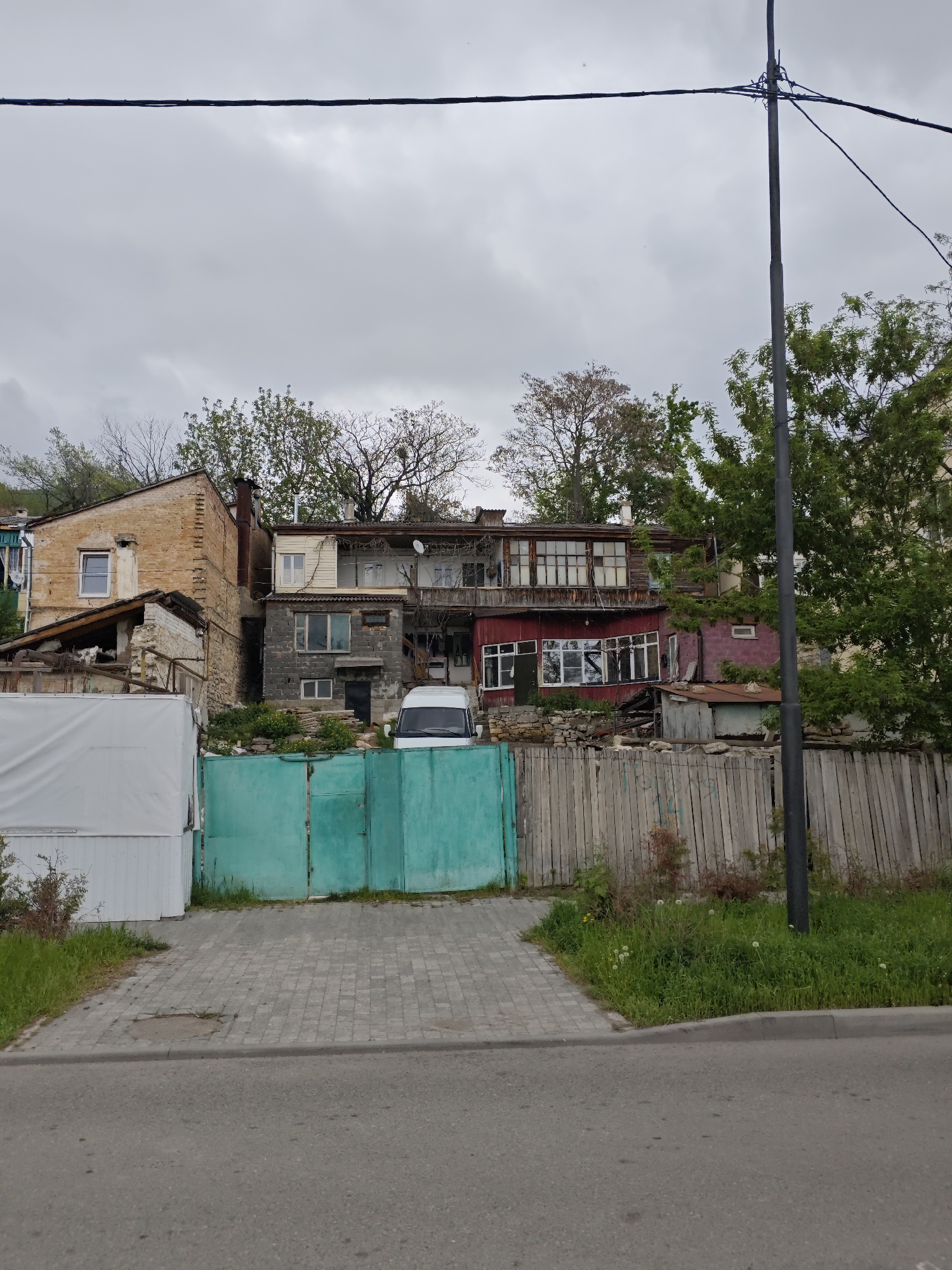 The photograph depicts a street view in what appears to be Pyatigorsk, Russia. The focus is on a collection of old, somewhat dilapidated buildings behind a green metal gate and a weathered wooden fence. A white van is visible parked behind the gate. The buildings are of varying construction and colors, including brick, wood, and painted surfaces. Some structures show signs of disrepair. Overgrown vegetation is scattered throughout the scene. A street and paved sidewalk are in the foreground. An electric pole is visible on the right. The sky is overcast.