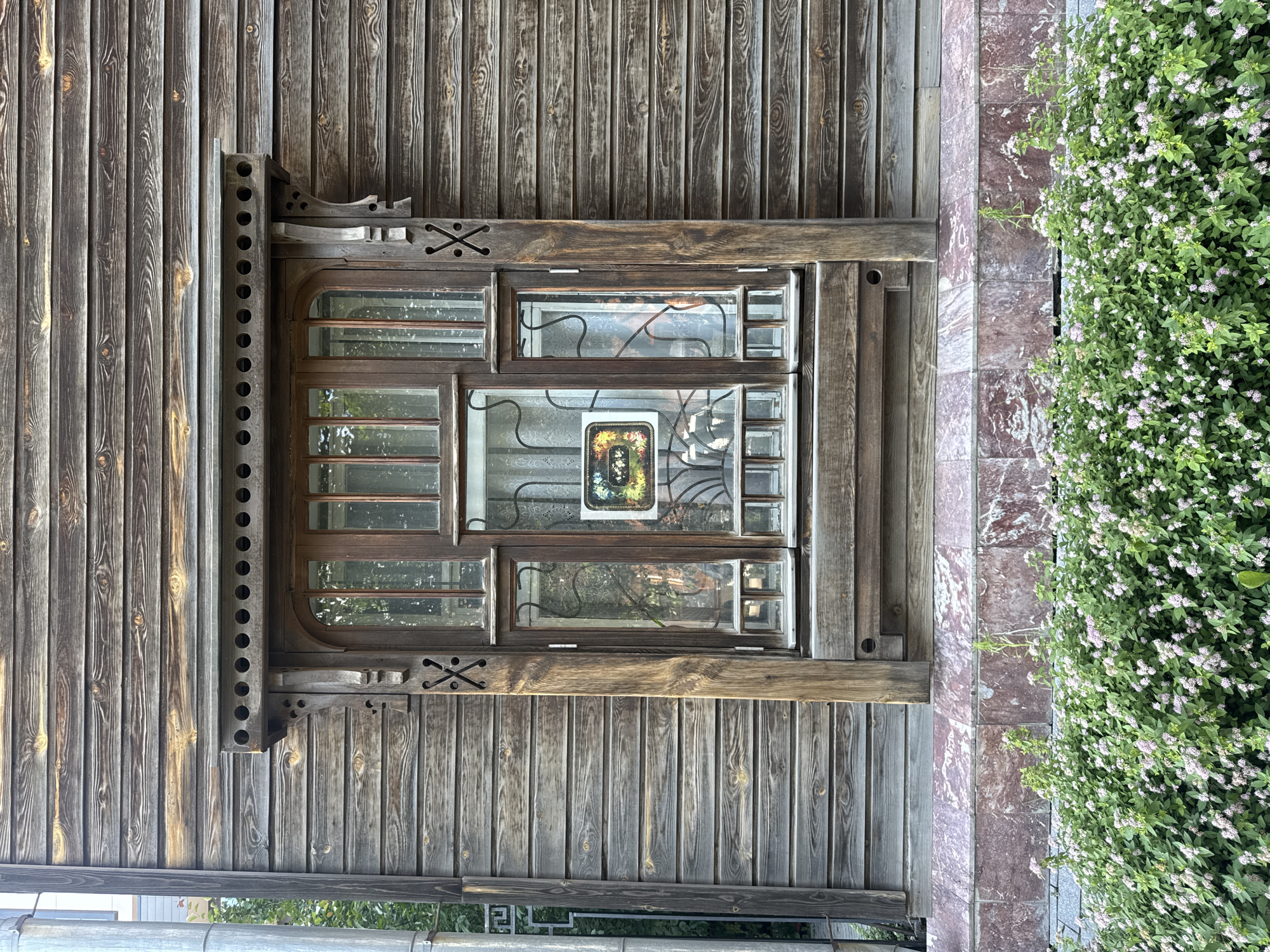 The image shows a close-up of an old wooden house with a detailed window in Yekaterinburg, Russia. The house is clad in horizontal wooden planks. Above the window, there is ornate wooden trim with decorative circular cutouts and brackets. The window itself is made of wood with multiple panes, some of which are obscured by a decorative metal grille. Inside the window, a small framed picture is visible. Below the window, there is a tiled base with pinkish-red marbled tiles. In the foreground, a dense bush with small white and pink flowers is blooming. The overall impression is of a rustic, possibly historic, building.