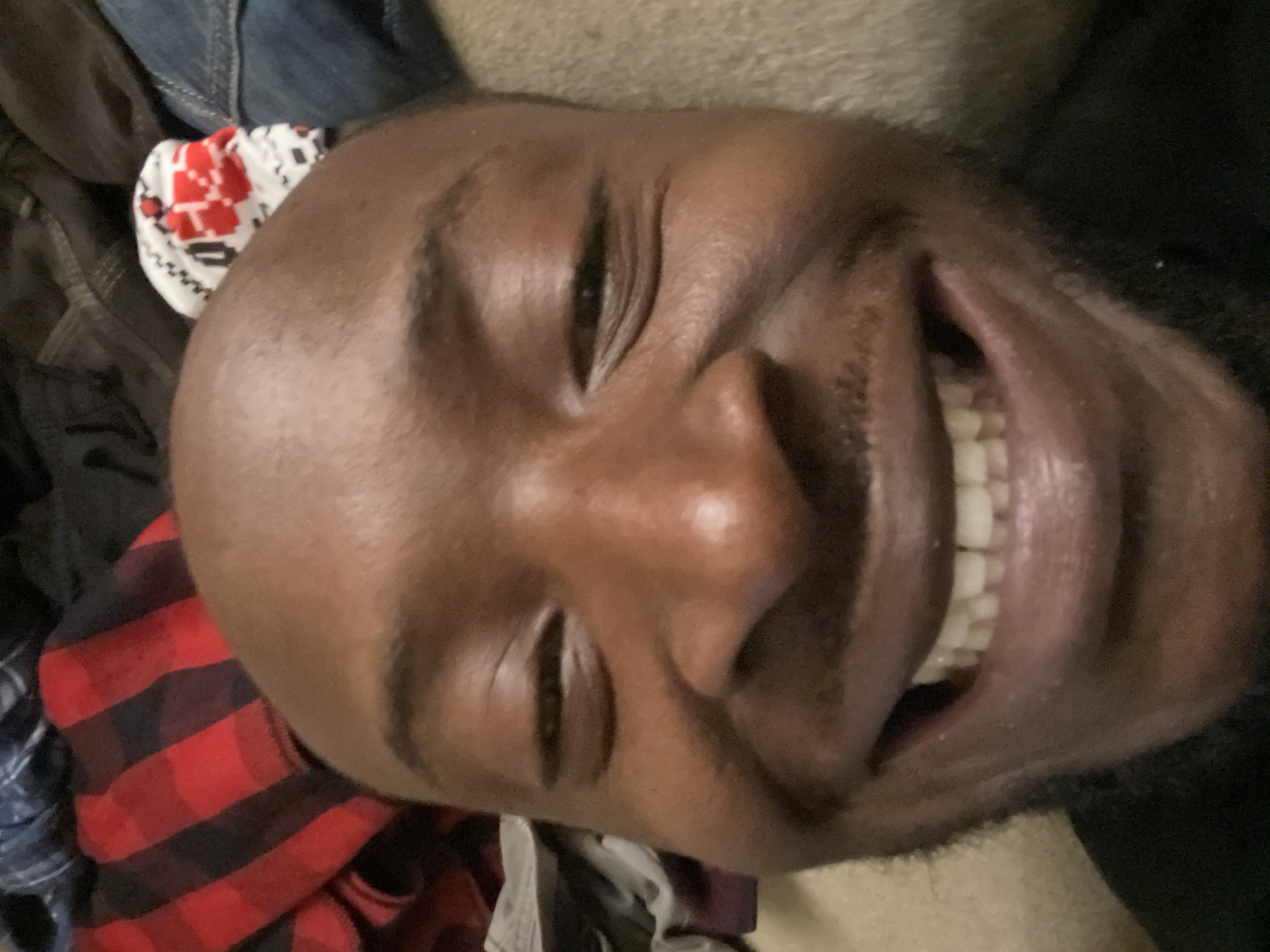 The image features a person, likely an adult male with dark skin, smiling broadly, with their head tilted to the side. The focus is on their face and upper torso.
**Subjects:** The primary subject is an individual, whose face shows a wide, joyful smile.
**Objects:** The person is wearing a red and black plaid shirt. Next to their head, a white item with red and black patterns, possibly a sock or a small knitted cap, is visible. Dark fabrics, resembling clothing or bedding, are also present in the background.
**Scene, Setting, and Location:** The scene appears to be indoors, suggested by the soft, neutral background which could be a bed, couch, or floor with textiles. The person is positioned horizontally or lying down. The location context provided is Jos, Nigeria.
**Activity or Event:** The person is laughing or exhibiting a very wide, cheerful smile. Their eyes are squinted or nearly closed due to the intensity of their smile, and their mouth is open, revealing their teeth.
**Notable Details:** The prevailing emotion is one of happiness, joy, or amusement. The lighting is soft and even, without strong shadows, typical of an indoor setting. There are no specific indicators for the time of day or weather within the image.
**Visible Text:** There is no visible text in the image.