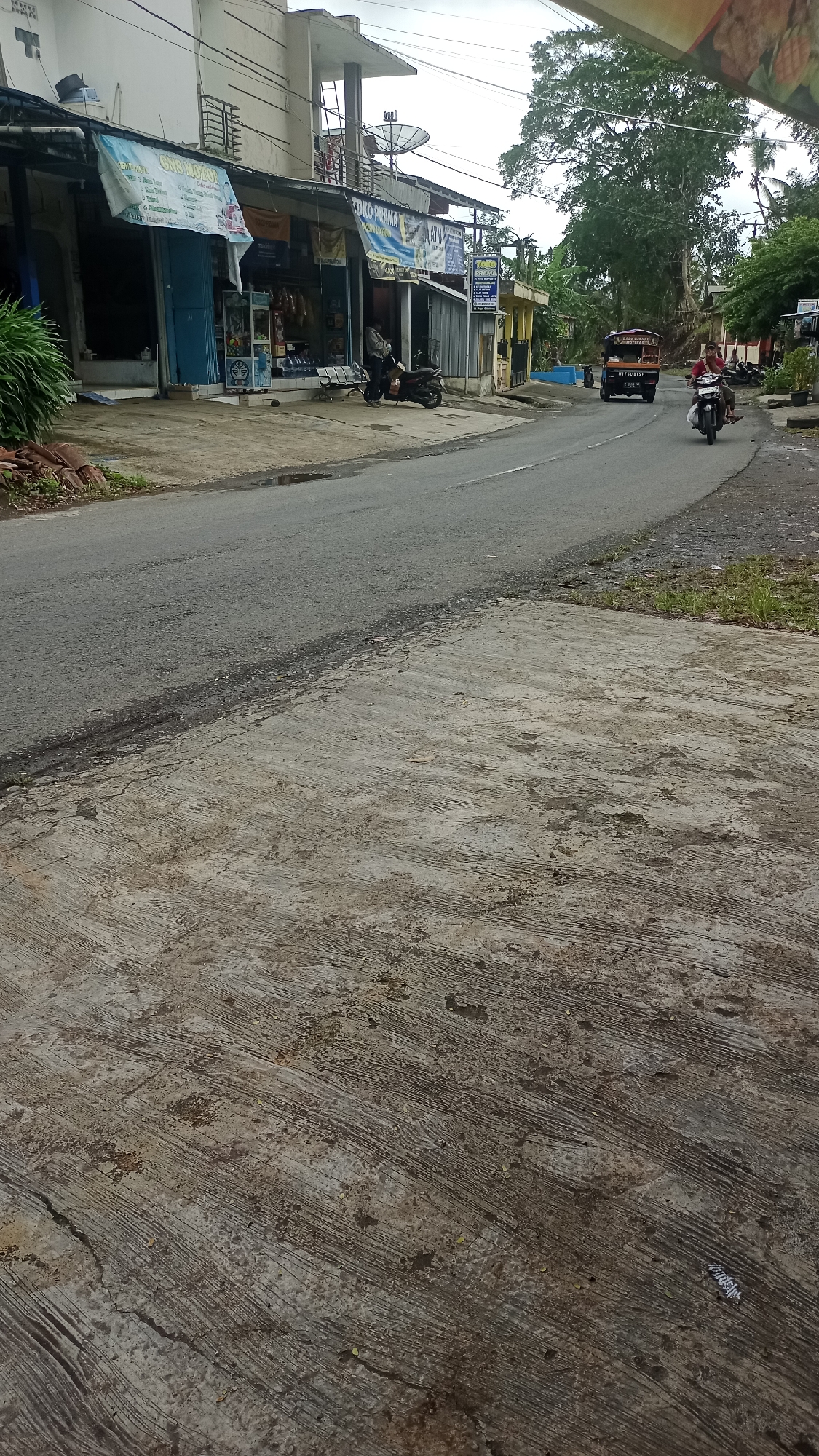 The image depicts a street scene in Karyamandala, Indonesia. On the left, a row of shops with awnings line the street. One shop has a blue door and displays various items inside, including bottles and what appear to be snacks. A motorbike is parked in front of this shop. Further down the street, another shop, "TOKO PRAMA," is visible, with signs indicating "ATM" and other services. A man in a gray jacket is seated on a motorbike in front of this shop. Across the street, two motorbikes are in motion. One, a red and black Mitsubishi truck, is traveling away from the viewer, and a rider on a black motorbike is following it. A man is riding the motorbike, wearing a red shirt. The road is paved and appears to be slightly wet in places, with some grass and debris on the sides. The background features a large tree with lush green foliage and a cloudy sky. The foreground consists of a concrete sidewalk with a rough, textured surface.