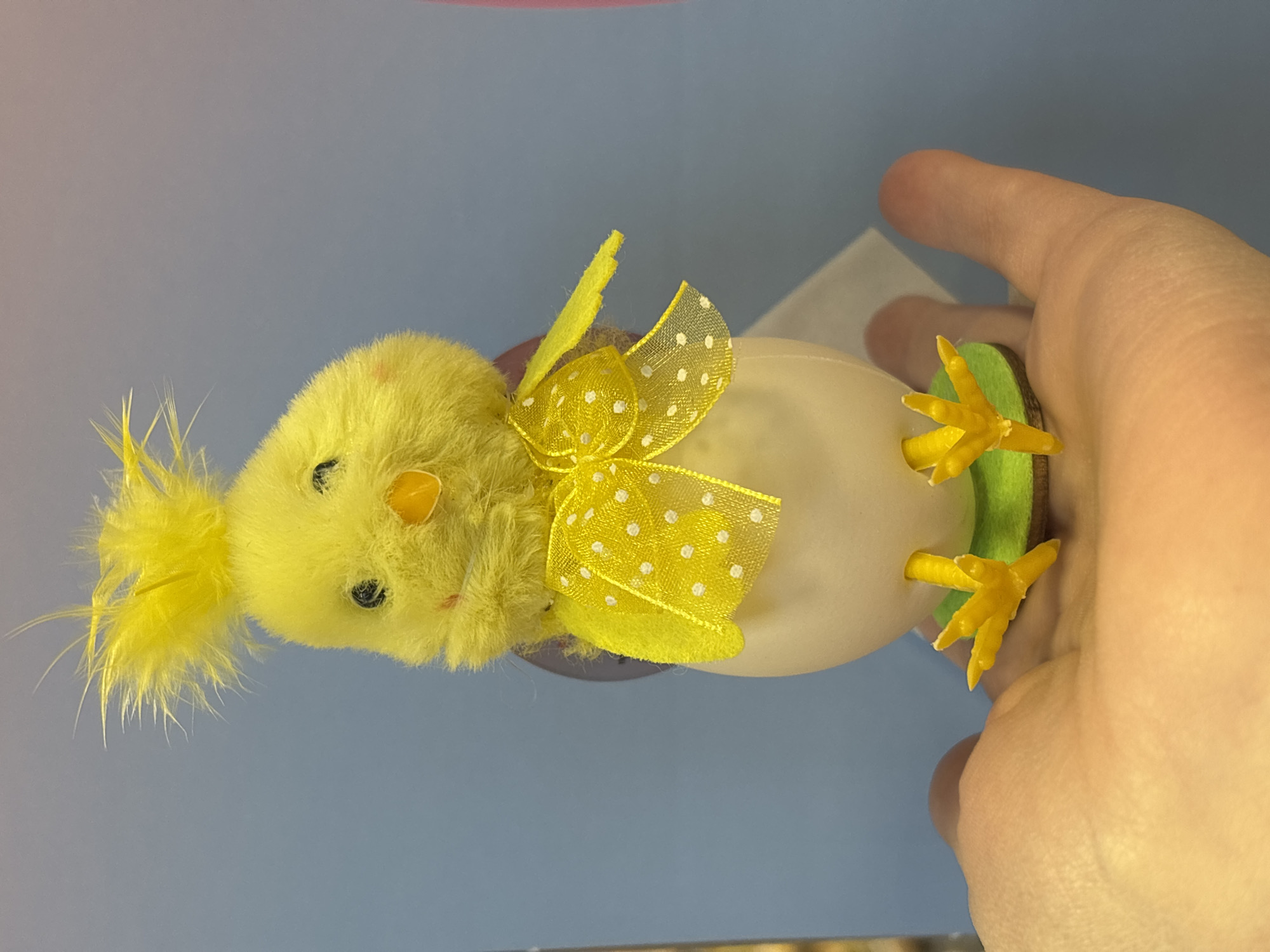 A yellow chick decoration with a dotted ribbon on its head sits atop a white egg-shaped object. The base of the object is green, with yellow chick feet attached. A hand is holding the decoration. The background is a gradient of blue and pink. There is no indication that this image was taken in Izhevsk, Russia.