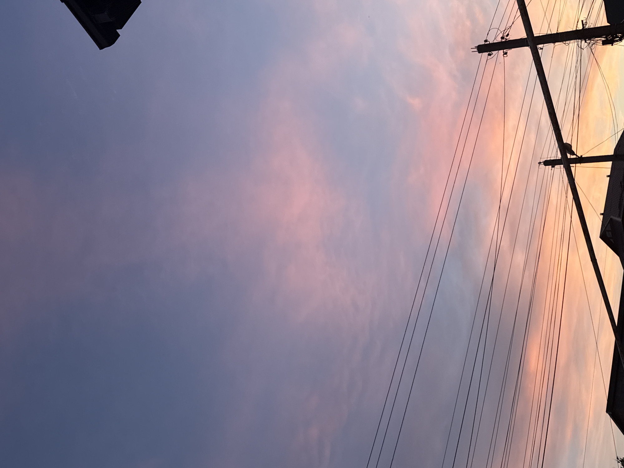 The image is an upward view towards the sky, which is colored with shades of pink, purple, and orange, likely during a sunset or sunrise. In the foreground, there are electrical poles and wires that crisscross the sky, slightly obscuring the view of the colorful clouds. Parts of buildings are visible on the periphery of the frame, both at the top left and the right edge. Given that the location is Kaspiysk, Russia, this is likely a residential or urban area with standard utility infrastructure. There are no people visible in the image. The photograph seems to capture a serene moment with a combination of natural beauty and urban elements.