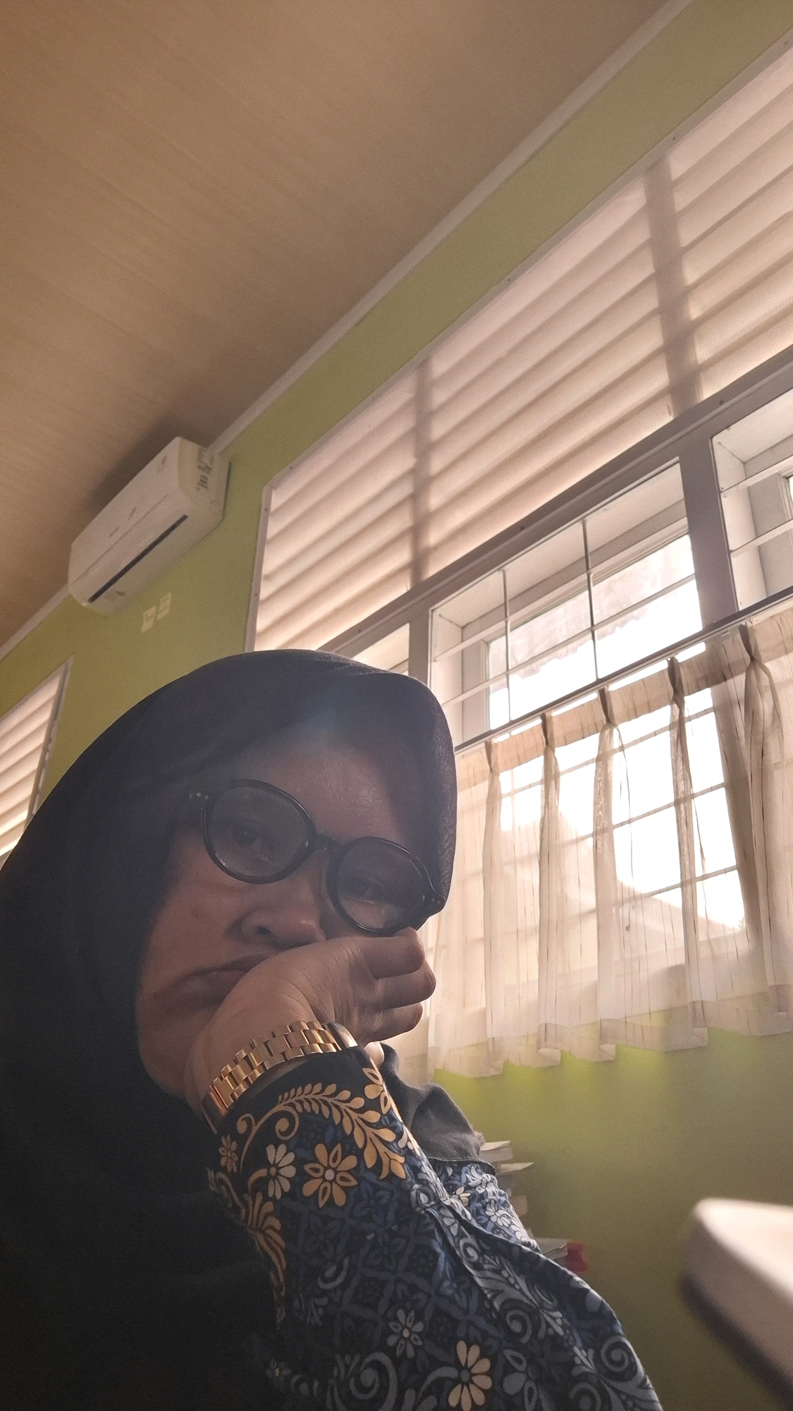 The image is a close-up selfie of a person wearing a black hijab and glasses. The person is resting their head on their hand, with their fingers touching their chin. They are wearing a patterned blue and gold shirt. The background shows a room with a light green wall, a wooden ceiling, and a large window with blinds and sheer curtains. An air conditioning unit is mounted on the wall. The scene appears to be indoors, possibly in a home or office, in the city of Bandar Lampung, Indonesia.