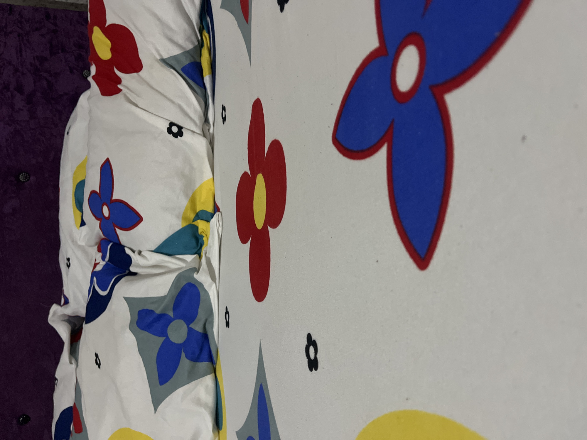 The image shows a close-up of a bed with a flower-patterned white comforter. The background includes a purple headboard. The location is unknown, and the prompt about Abuja, Nigeria, is irrelevant.