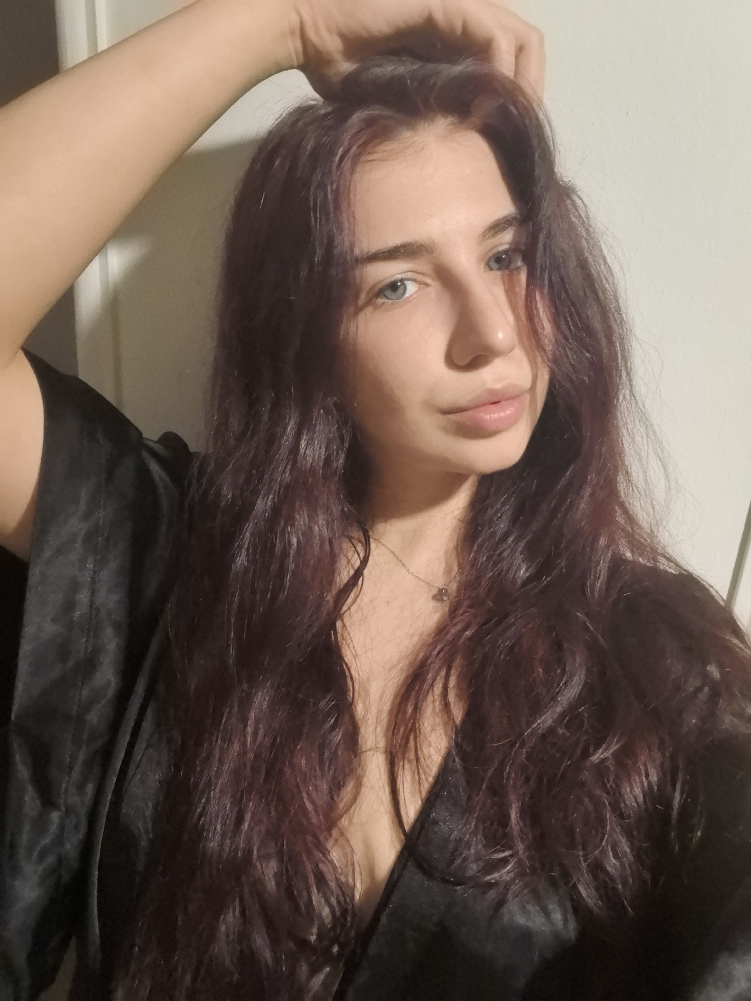 A medium shot depicts a woman with light skin, blue eyes, and long, wavy, dark red hair. She is wearing a black silky robe or garment, and her right arm is raised with her hand touching her hair. A thin necklace is visible around her neck. The background is a plain, light-colored wall. The location is Unknown, Unknown.