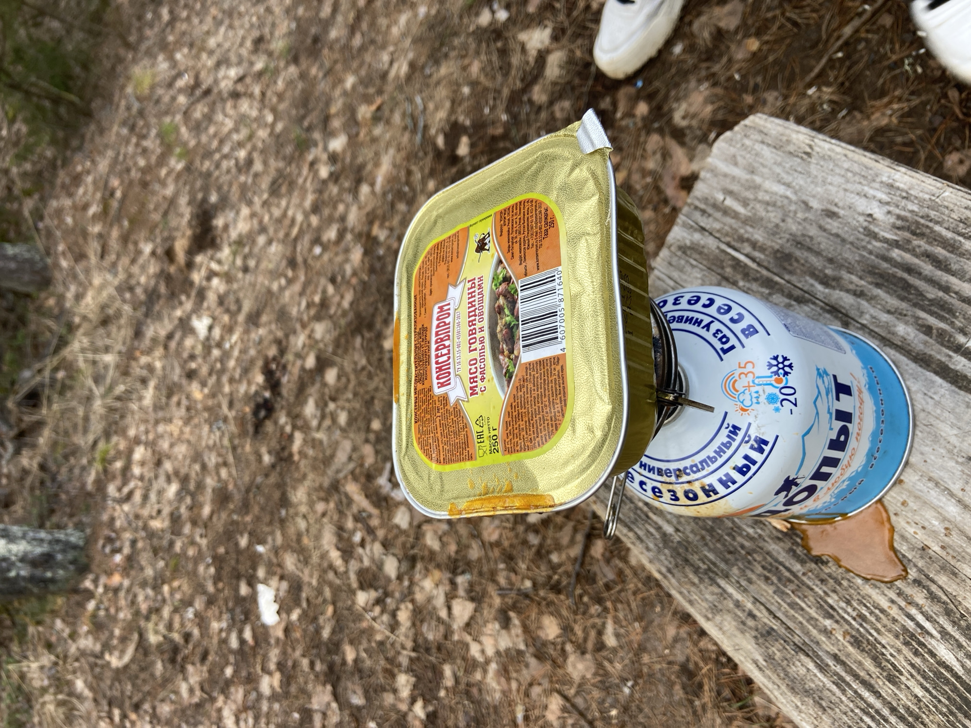 The scene presents a still life of camping provisions on a weathered wooden surface, likely a rough-hewn table or log. The focus is on a square, golden-colored tin can of "Meat with Beans and Vegetables" featuring Cyrillic text and the brand "Konservprom," indicating a Russian origin. Next to the can is a gas canister of "Universal" gas, implying it is suitable for all seasons and can be used in temperatures as low as -20 degrees Celsius. A brown liquid, possibly grease or sauce from the food, has spilled onto the wooden surface near the canister. The backdrop reveals a forest floor covered in dry leaves and pine needles. Parts of two white sneakers are visible in the upper part of the image. It seems that the person wearing the sneakers is either preparing a meal, having already cooked one, or is about to leave from the site. Given the Cyrillic text, it could potentially be the city of Chaikovsky, Russia.