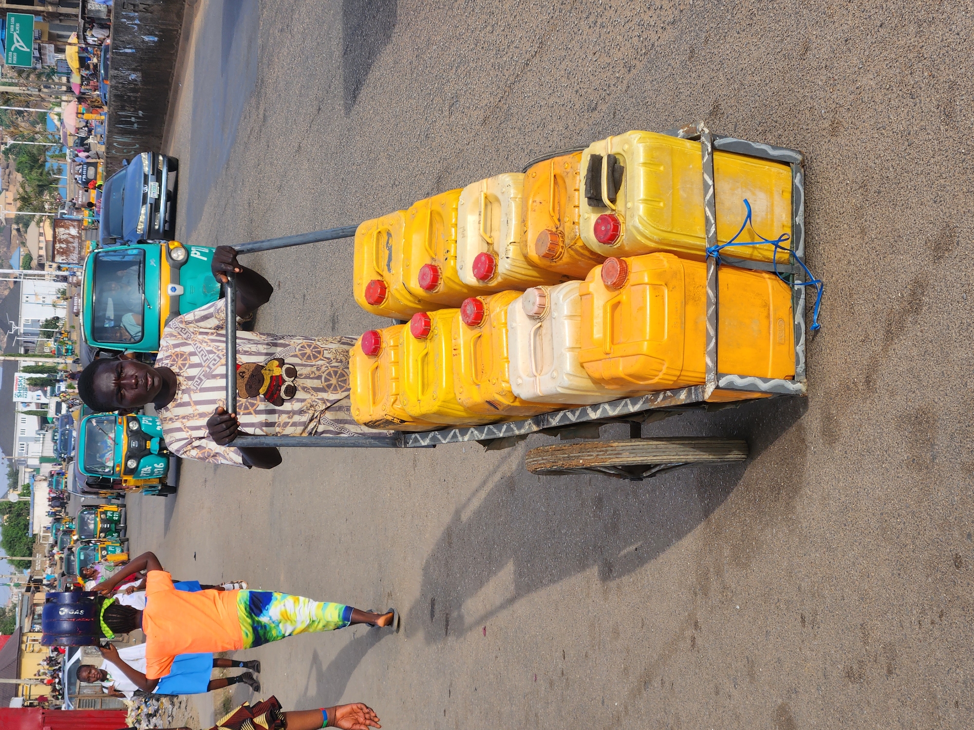 A man is pulling a handcart loaded with yellow plastic jerrycans along a street in what appears to be the city of Jos, Nigeria. The jerrycans are arranged in two rows, and secured to the cart with blue string. The man is wearing a patterned shirt. In the background, there are several auto-rickshaws (tuk-tuks) and cars parked along the side of the street. Other individuals are walking on the roadside. The street is paved.