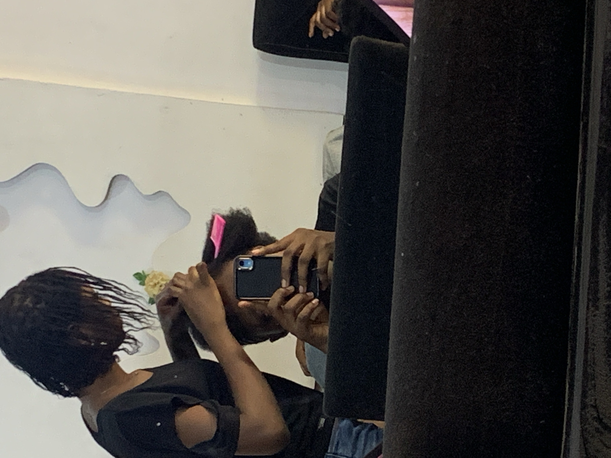 A woman stands in front of a mirror, holding up a cell phone to photograph her reflection in a brightly lit room. The woman has short, dark, curly hair with a pink barrette. She wears a dark-colored, short-sleeved top and blue jeans. In the reflection, a small yellow flower is near her head. The background behind the woman and her reflection is a white wall with a grey cloud-like design. In the foreground, partially obscuring the view, are two dark, cylindrical objects. There is no definitive evidence to confirm this image was taken in Jos, Nigeria.