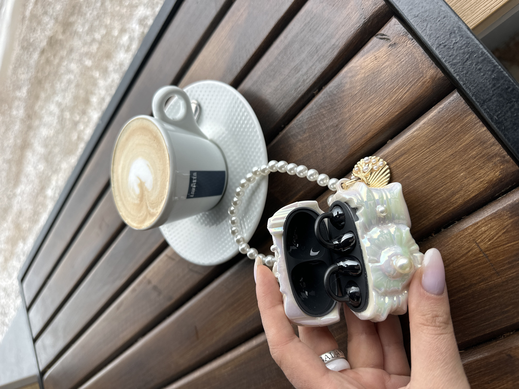 The scene is set on a wooden slat table, likely in an outdoor setting in Šušanj, Montenegro, based on the provided information. A cup of coffee sits on a white saucer to the left. To the right, a hand with manicured nails holds an open, iridescent white seashell-shaped case containing black wireless earbuds. The case is adorned with a string of pearls and a small golden charm. The hand also has a silver ring on the ring finger. The location appears to be a café or similar establishment in Šušanj.