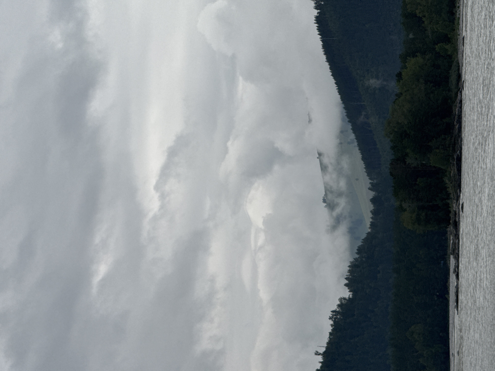 The image depicts a cloudy, overcast day over a river and forested hills. The foreground features a wide, choppy river with several dark rocks visible near the banks. Beyond the river, a dense, dark green forest covers the lower slopes of the hills. The hills in the middle ground are partially obscured by thick, white clouds that weave through the landscape, obscuring higher peaks. The sky is filled with heavy, grey clouds, with some lighter patches suggesting diffused sunlight. There are no people or man-made structures clearly visible in the frame. The scene conveys a sense of natural, rugged terrain under a somber sky. The location is identified as Nizhnekayancha, Russia.