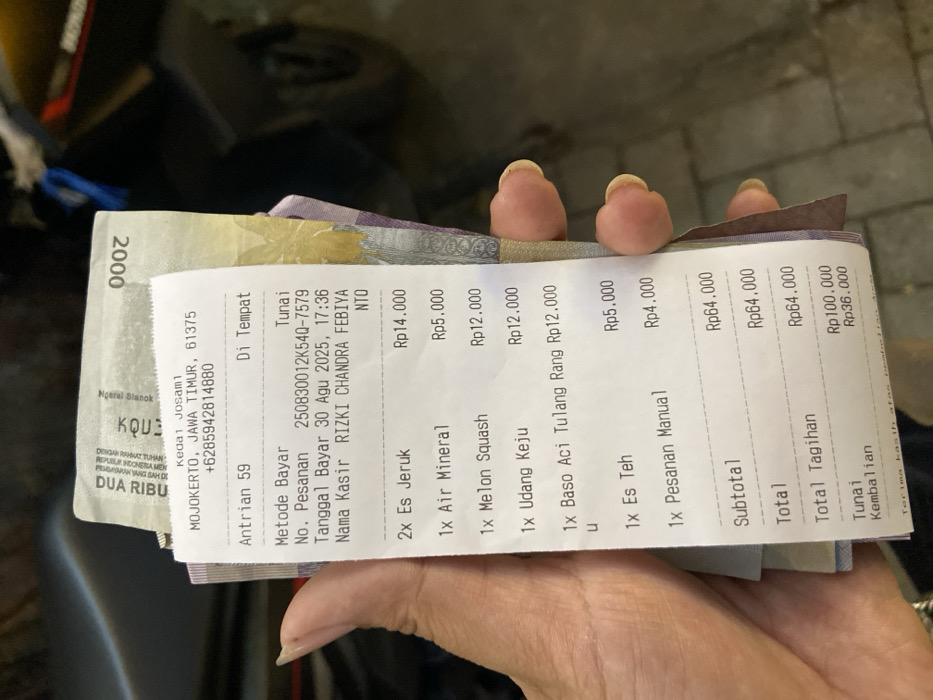 A close-up, slightly overhead view captures a human left hand holding a white paper transaction receipt and a stack of Indonesian Rupiah banknotes. The hand displays manicured fingernails, with some visible residue beneath the nail tips. The receipt, issued by "KEDAI Josam1," identifies its location as Mojokerto, Jawa Timur, with a postal code of 61375 and a contact number. The transaction occurred on August 30, 2025, at 17:36, for order number 59, paid by cash, and processed by cashier RIZKI CHANDRA FEBIYA. The itemized bill includes two Es Jeruk (Rp14.000), one Air Mineral (Rp5.000), one Melon Squash (Rp12.000), one Udang Keju (Rp12.000), one Baso Aci Tulang Rang (Rp12.000), one Es Teh (Rp5.000), and one Pesanan Manual (Rp4.000), totaling Rp64.000. A cash payment of Rp100.000 resulted in Rp36.000 change. Several Indonesian Rupiah banknotes are partially visible behind the receipt, with a Rp2.000 note clearly identifiable. The background is dark and out of focus, presenting indistinct shapes and textures, likely an interior or dimly lit exterior setting. The location specified on the receipt is Mojokerto, Jawa Timur, not Cembor.