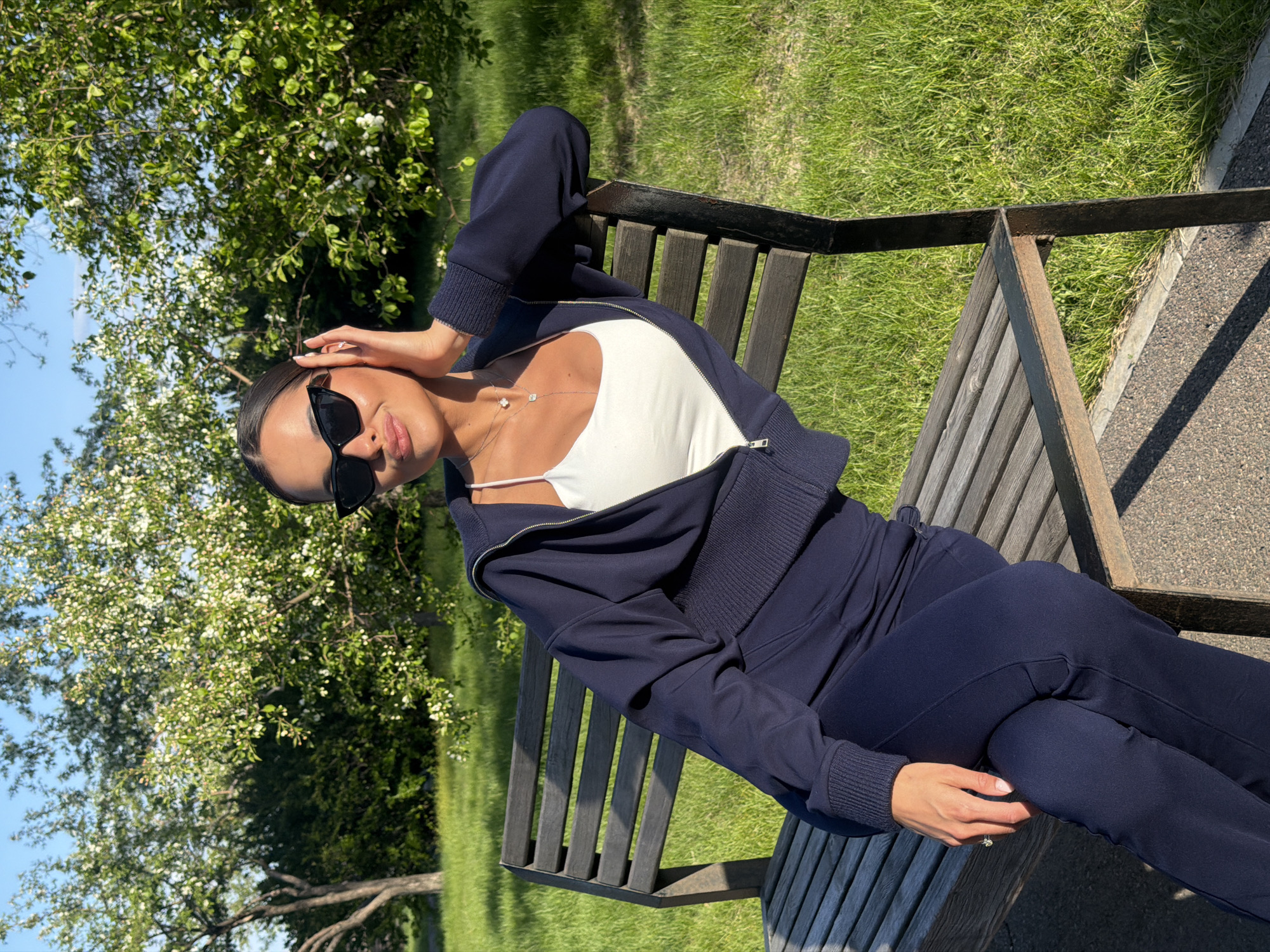A woman is sitting on a wooden bench in a park, wearing dark sunglasses, a white tank top under a partially zipped navy blue hooded sweatshirt, and matching pants. She has one hand on her head. The location appears to be an outdoor park area during daylight, possibly in Krasnoyarsk, Russia, as suggested by the prompt. There is grass, a tree with white blossoms, and a paved walkway.