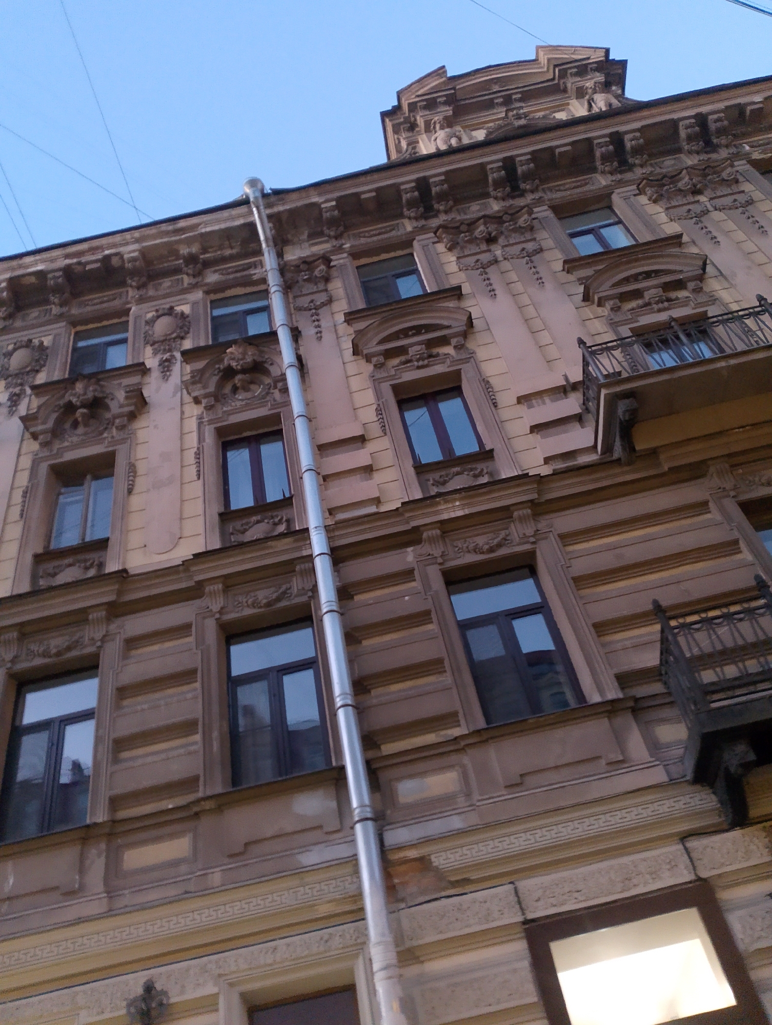 This is a low-angle, full-shot photograph of the facade of a historic building in Saint Petersburg, Russia. The building features ornate architectural details, including decorative carvings and moldings, common in the city's classical and baroque styles. Several windows of various sizes and shapes punctuate the facade. A prominent metal downspout runs vertically along the building's side, suggesting it is a functional element of the building's drainage system. The sky above is clear and pale blue, indicating it is likely daytime or dusk. The image appears to be taken from a street or courtyard looking upwards.