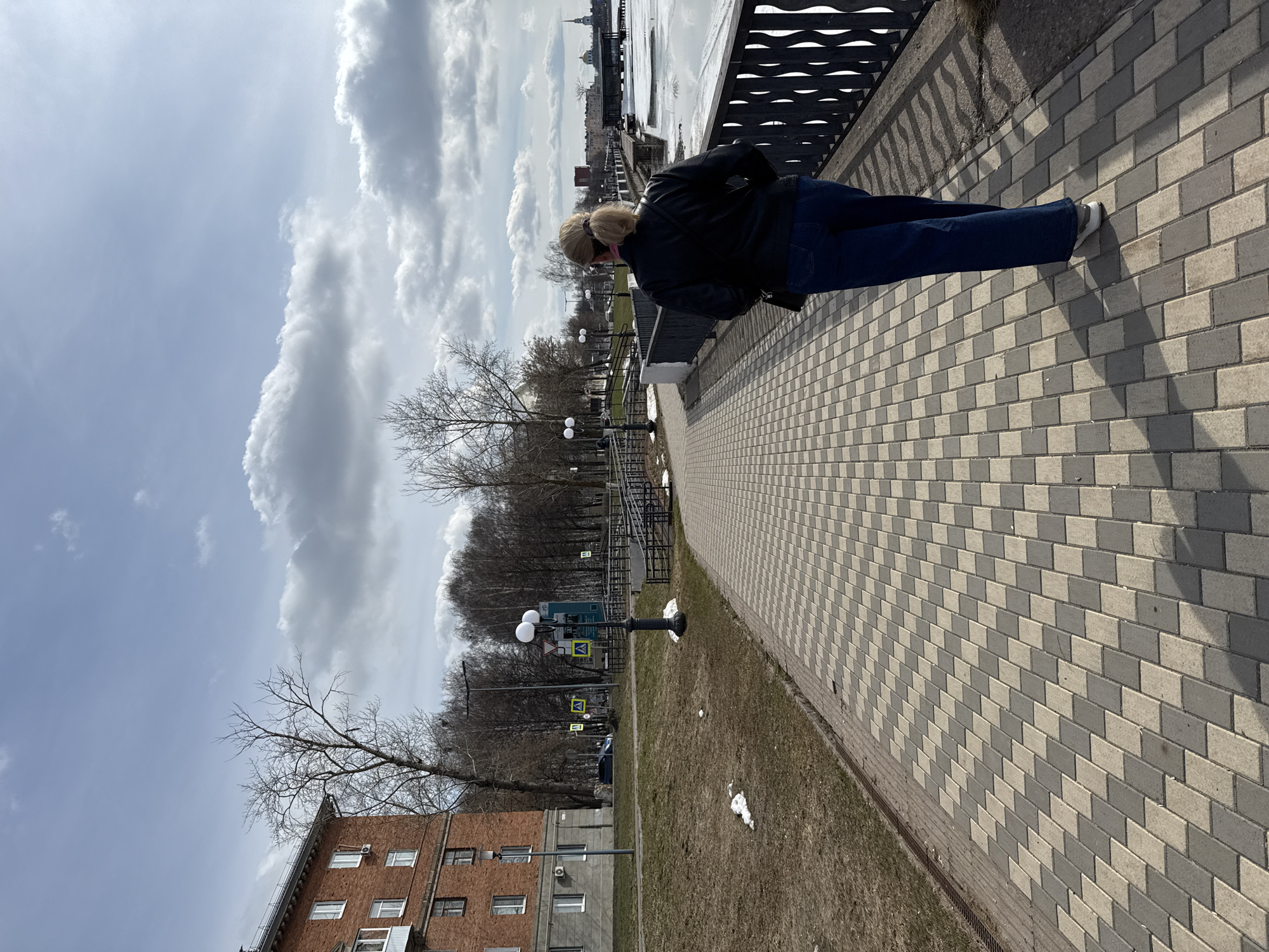 A woman wearing a black jacket and blue jeans stands on a brick walkway in Votkinsk, Russia. She is facing a metal railing next to a body of water, presumably a river or pond, which is partially frozen. In the background, buildings and trees are visible under a cloudy sky. Street lamps are lined along a path beside the walkway. A brick building is visible to the left. The walkway itself is made of light and dark colored bricks in a patterned design.