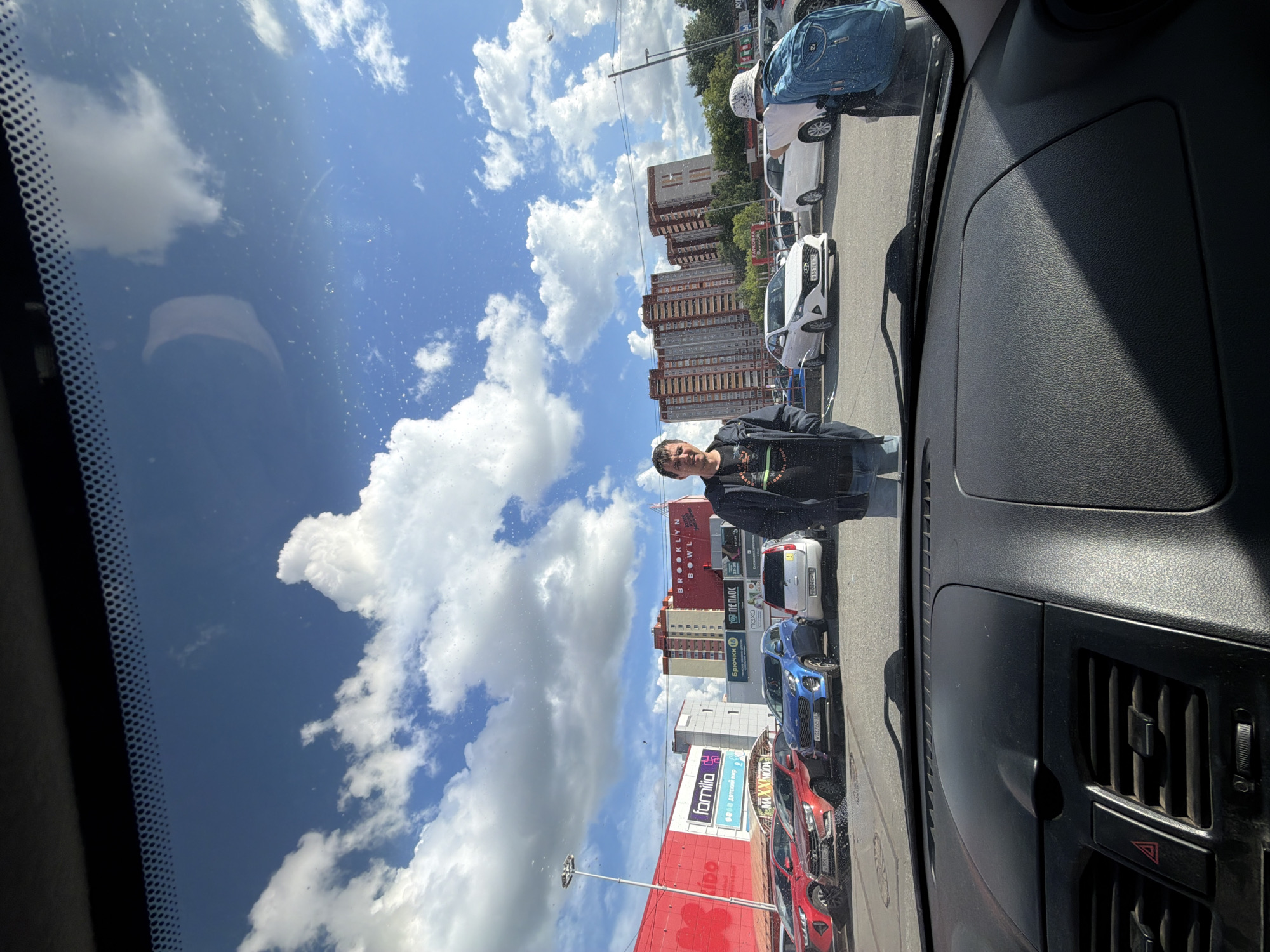 The image, taken from inside a car, shows a view of a street scene in Izhevsk, Russia, near the "Familia" store. A man wearing a black jacket and a light-colored hat stands on the street near several parked cars. Buildings are visible in the background under a partly cloudy sky. The car's dashboard and windshield partially frame the view.