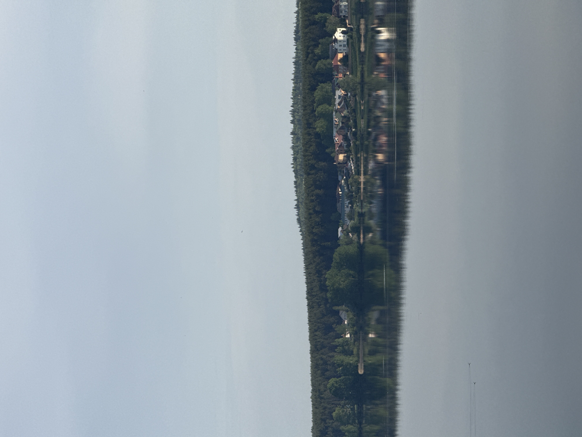 The image depicts a body of water reflecting a treeline and buildings. It's a view of a landscape, where the reflection appears as an extension of the real world. Two distant vertical objects, possibly boats or structures, are visible on the water. The top part shows a hazy sky. The scene appears to be set in Votkinsk, Russia, due to the type of landscape and architecture.