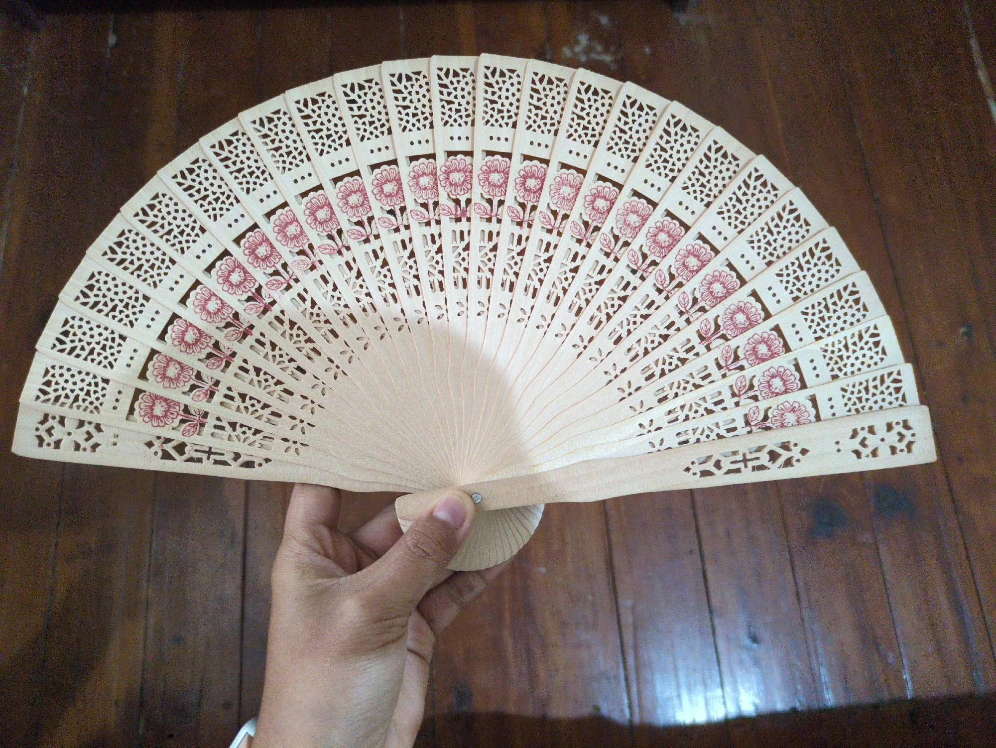 A hand holds an open, intricately carved wooden fan adorned with pink floral patterns. The fan is fanned out on a wooden surface, appearing to be a floor or table, with visible wood grain. The background is a dark, textured wooden surface. The image is a top-down view. The setting is not specified to be in the city of Bhagsu, India.