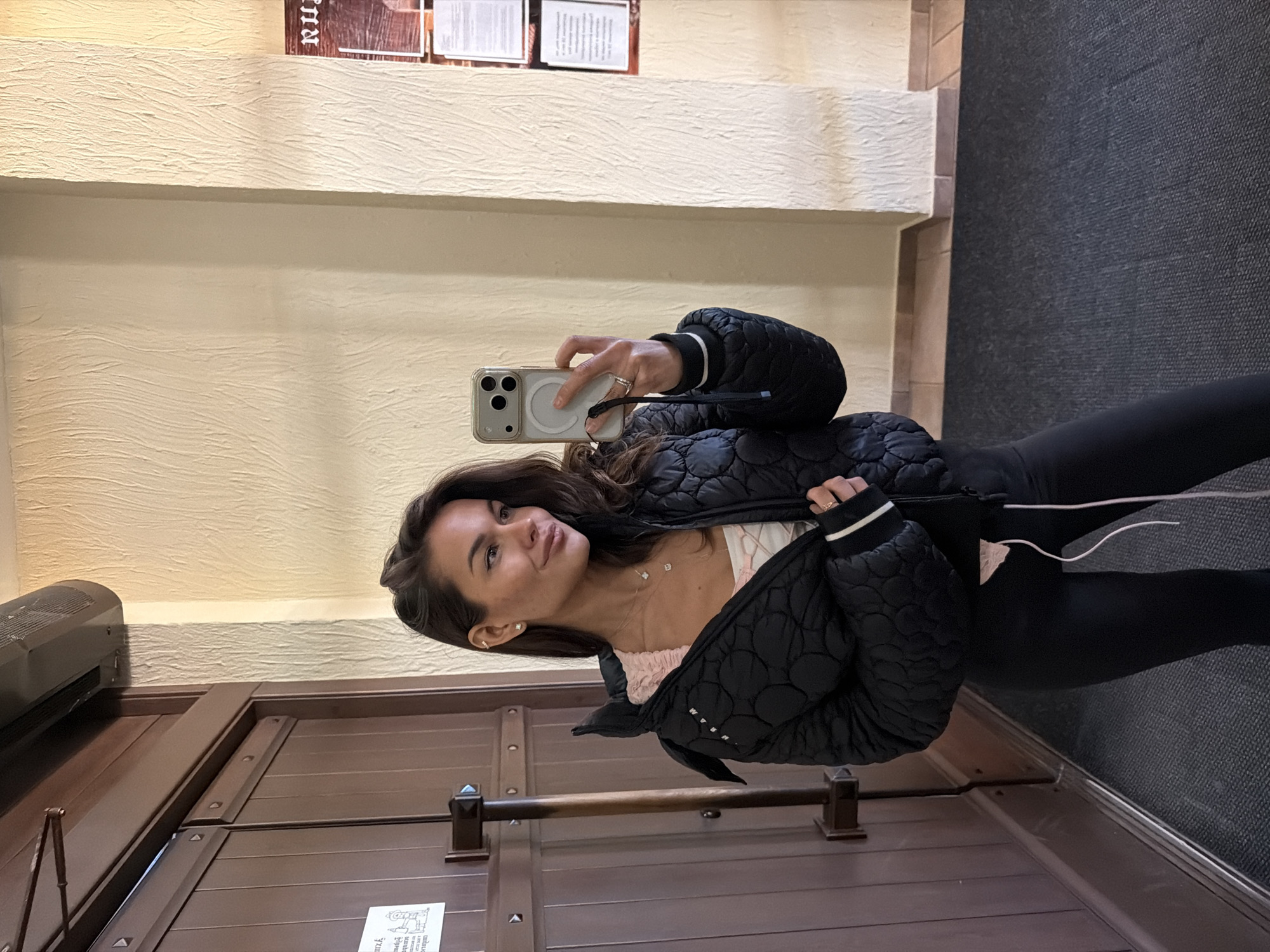 A woman is taking a selfie indoors. She is wearing a black quilted jacket and black leggings. She is holding a smartphone up to her face. The background includes a textured beige wall, a dark wood door with metal hardware, and a section of gray carpet. Two framed documents are visible on the wall. A wall-mounted air conditioner is partially visible above the door. The location is most likely not Krasnoyarsk, Russia as the scene seems more indicative of an indoor location than a specific recognizable city.