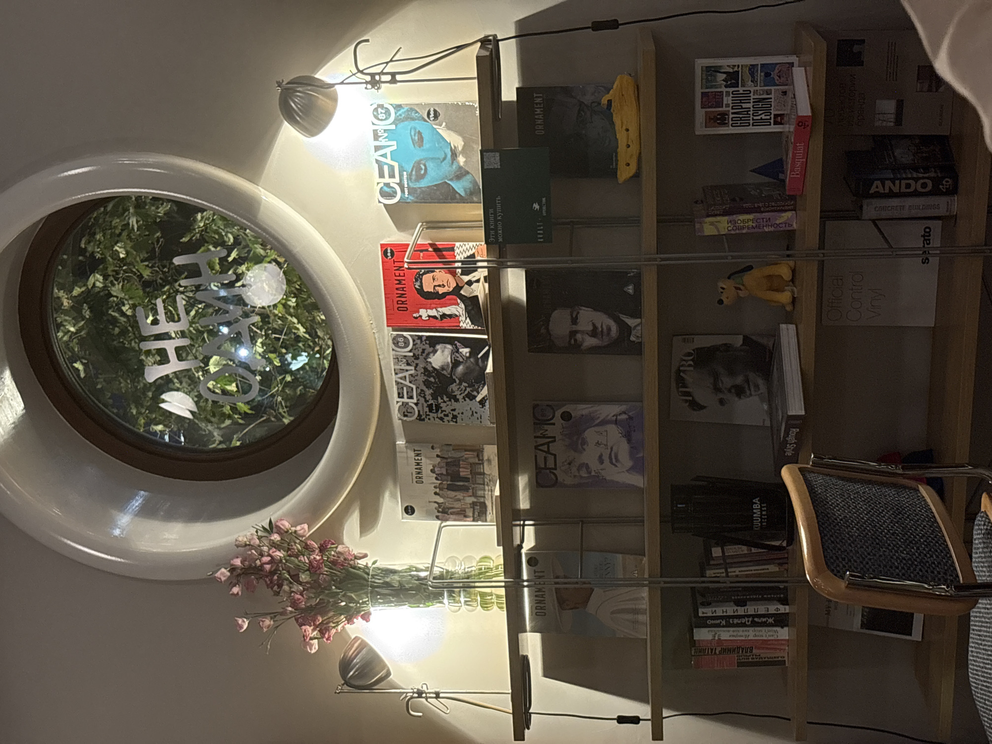 The image depicts an interior scene, likely a study or living room, featuring a wooden shelf unit displaying various books, magazines, and decorative items. A circular window with greenery visible through it is positioned above the shelves. A vintage-style lamp illuminates one of the shelves, where several magazines with "ORNAMENT" and "CEAMO" titles are displayed, alongside books featuring portraits and artistic designs. Below, more books are organized, including titles like "Graphic Design," "Basquiat," and "Ando." A plush toy and a small yellow submarine figurine also rest on the shelves. A chair with a woven seat is partially visible in the foreground. The overall ambiance is cozy and suggests a space dedicated to art, literature, and possibly music, given the magazine covers. The location of Rostov-on-Don, Russia, is not evident from the visual information within the image.