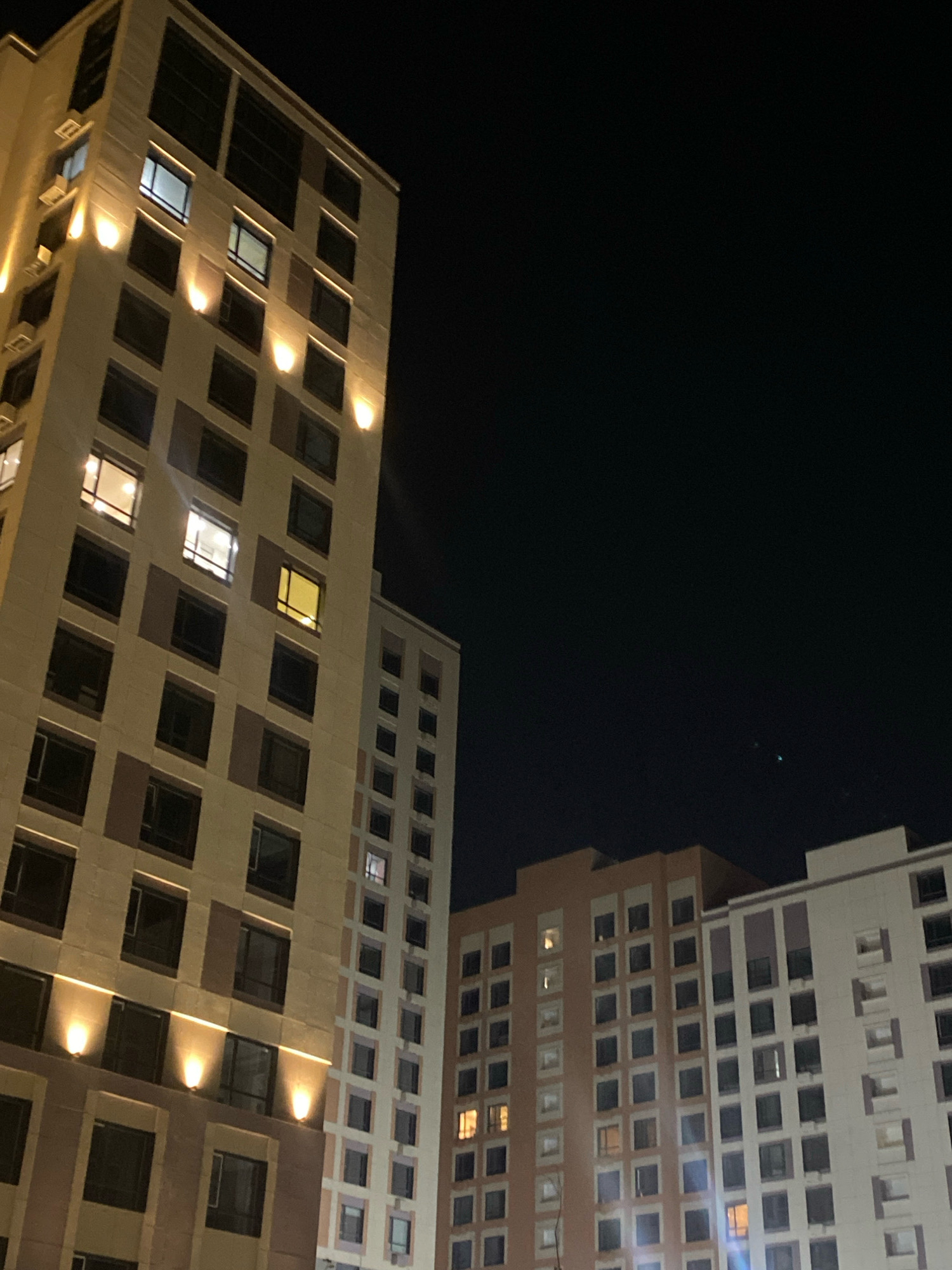 The image depicts a nighttime view of modern buildings in Astana, Kazakhstan. The foreground features a tall, light-colored building with numerous windows, some of which are illuminated with warm light, suggesting occupancy. Decorative yellow lights are strategically placed along the facade, casting a glow on the building's exterior. To the right, two other buildings are partially visible; one appears to be a similar height and architectural style, while the other, further in the background, is lighter in color and also has many windows. The sky is dark, indicating it is late evening or night, with a few faint celestial lights visible. The overall impression is of a well-lit urban environment at night.