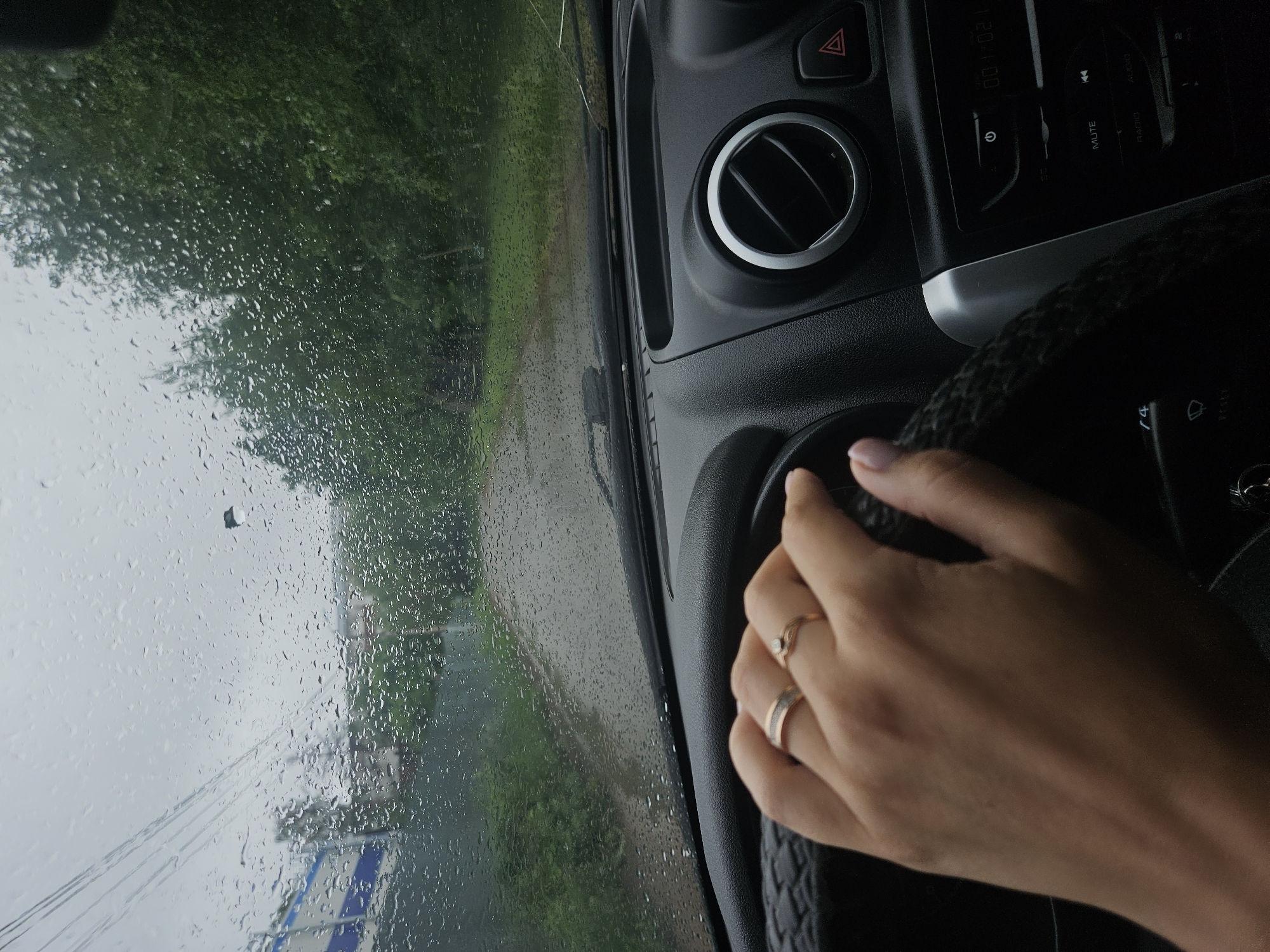 The image shows the interior of a car on a rainy day. The windshield is covered in raindrops. A hand with two rings on the ring finger is visible, resting on the steering wheel. Part of the dashboard is in view, including an air vent and a car stereo system. The background visible through the windshield depicts a road and greenery, suggesting an outdoor environment. Given the context of Chaikovsky, Russia, the location could be a road within the city or its surrounding areas, experiencing rainfall.